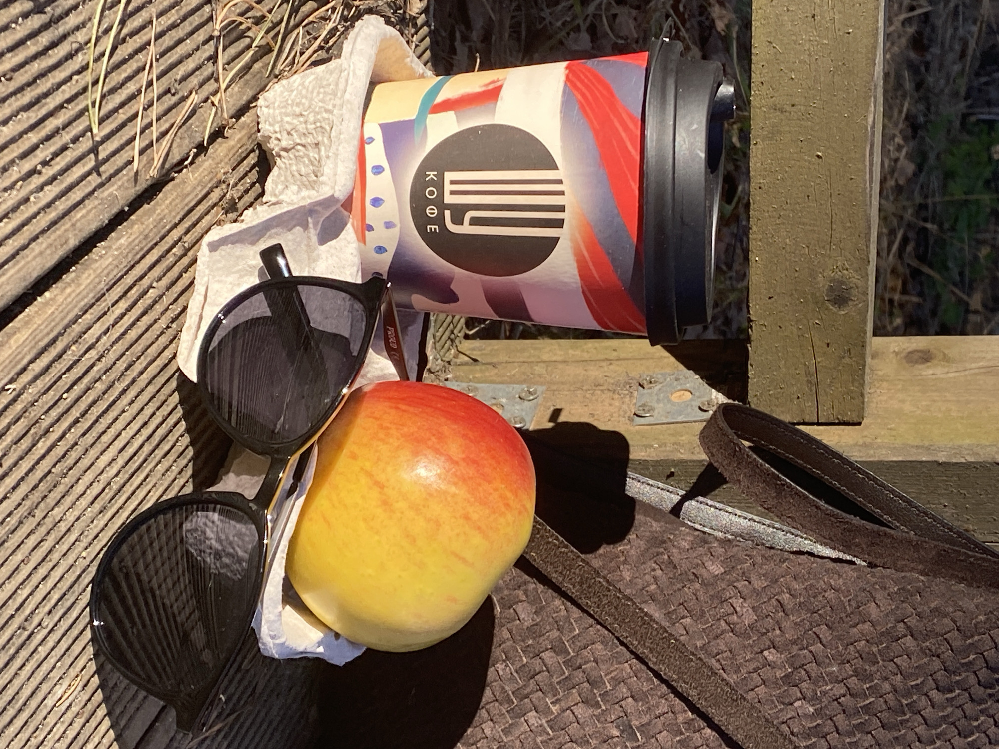 The image depicts a still life arrangement outdoors, possibly in Chaikovsky, Russia, given the Cyrillic script "KOFE" (meaning coffee) on the cup. An apple, predominantly yellow with red highlights, rests near a pair of dark-framed sunglasses on a crumpled white tissue or napkin. A paper coffee cup with a black lid and red, yellow, and blue patterns is positioned behind these items. The items are resting on a weathered wooden surface with linear grooves, likely part of a bench or platform. A brown woven bag with leather straps is situated nearby. Sunlight casts shadows across the scene.