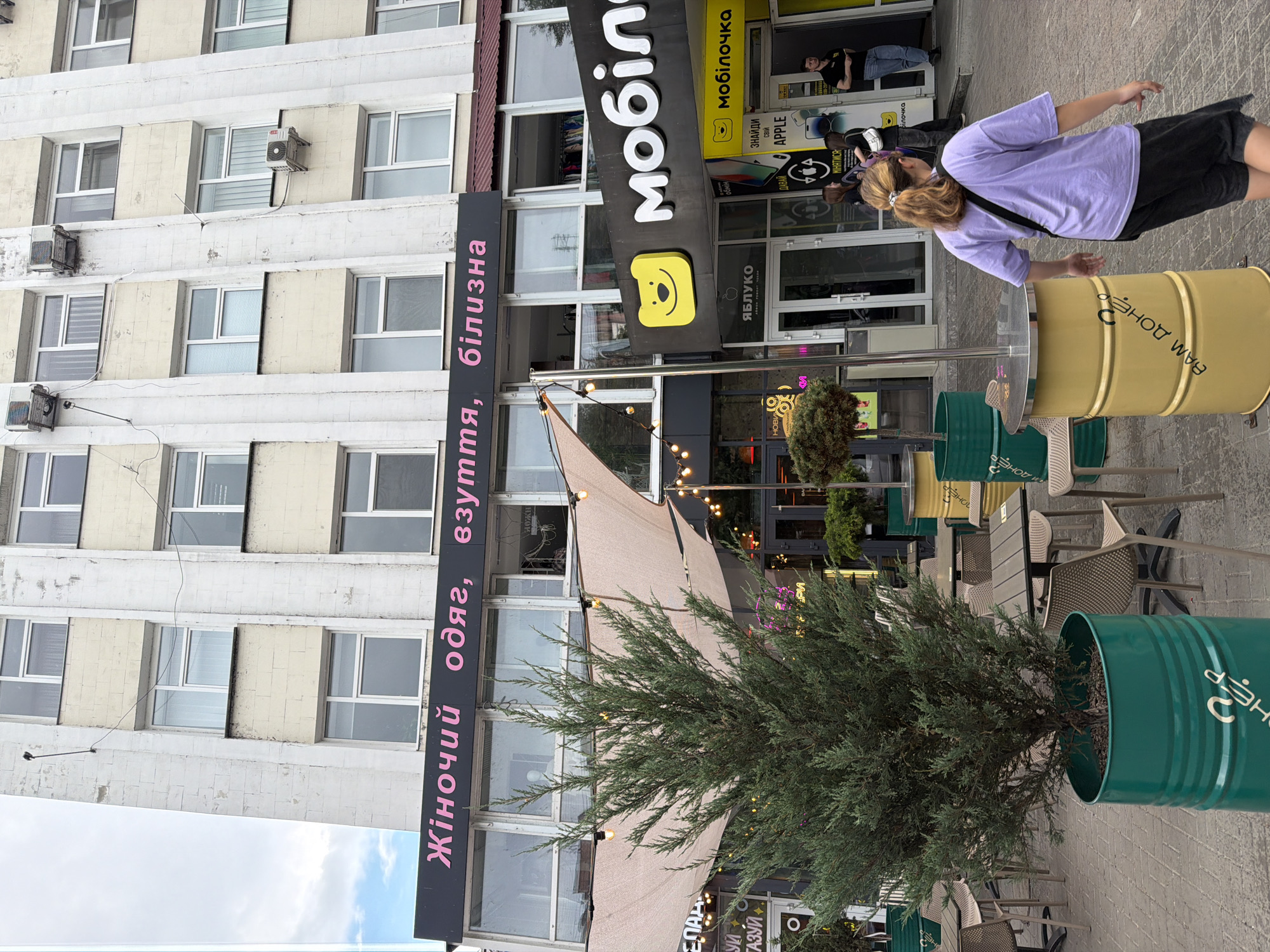 The image shows a street scene in Cherkasy, Ukraine, with a multi-story building in the background. A storefront with pink lettering reading "Жіночий одяг, взуття, білизна" (Women's clothing, shoes, underwear) is visible on the first floor. Next to it, a shop with a yellow smiling bear logo and the word "Мобілочка" is present. In the foreground, a woman with blonde hair in a ponytail, wearing a purple t-shirt and black shorts, is walking away from the camera. She has a black crossbody bag. Several outdoor tables and chairs are set up, along with large green and yellow cylindrical planters. The overall environment appears to be a commercial area.