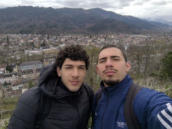 The image depicts two adult males in a close-up selfie taken from an elevated viewpoint overlooking the city of Freiburg im Breisgau, Germany. This vantage point is consistent with Schlossberg Hill, providing a panoramic view of the urban area nestled in a valley with forested mountains beyond.
The male on the left has dark, curly hair and a faint mustache, wearing a black hooded puffer jacket over a dark high-neck garment. A black backpack strap is visible on his right shoulder. The male on the right has short dark hair and a trimmed goatee and mustache, wearing a dark blue zip-up athletic jacket with white stripes on the right sleeve and a light-colored logo on the left chest. A black backpack strap is visible on his left shoulder. Both individuals are positioned side-by-side, looking directly at the camera with neutral expressions.
The background shows a dense urban landscape of Freiburg im Breisgau, composed of numerous buildings with pitched roofs, many featuring red or brown tiles, and some lighter-colored, modern structures. Beyond the city, a range of mountains covered in dense forest rises under an overcast, grey sky. In the immediate foreground, to the right, green foliage with white blossoms is visible.