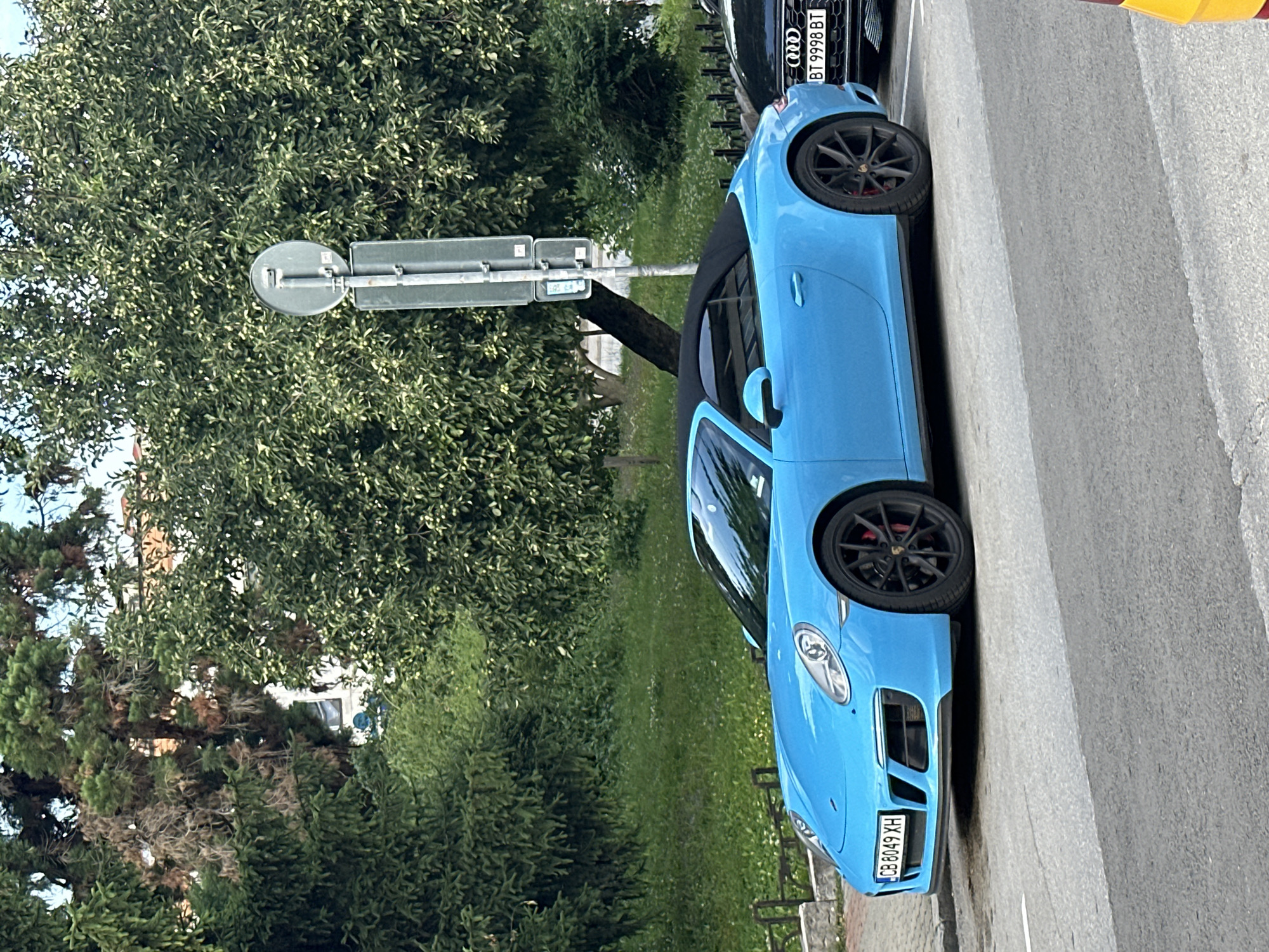 A bright blue Porsche convertible is parked on a city street in Veliko Tarnovo, Bulgaria. The car is facing left and has its black soft-top up. A black Audi is parked to the right of the Porsche, partially visible. The street is paved, and a sidewalk is visible in front of the car. Behind the cars, there is a grassy area with scattered white flowers, a low decorative fence, and several large, leafy trees. A tall signpost with a circular sign at the top is also present behind the Porsche. The license plate of the Porsche reads "CB 8049 XH", and the license plate of the Audi reads "BT 9998 BT". The sky is overcast.