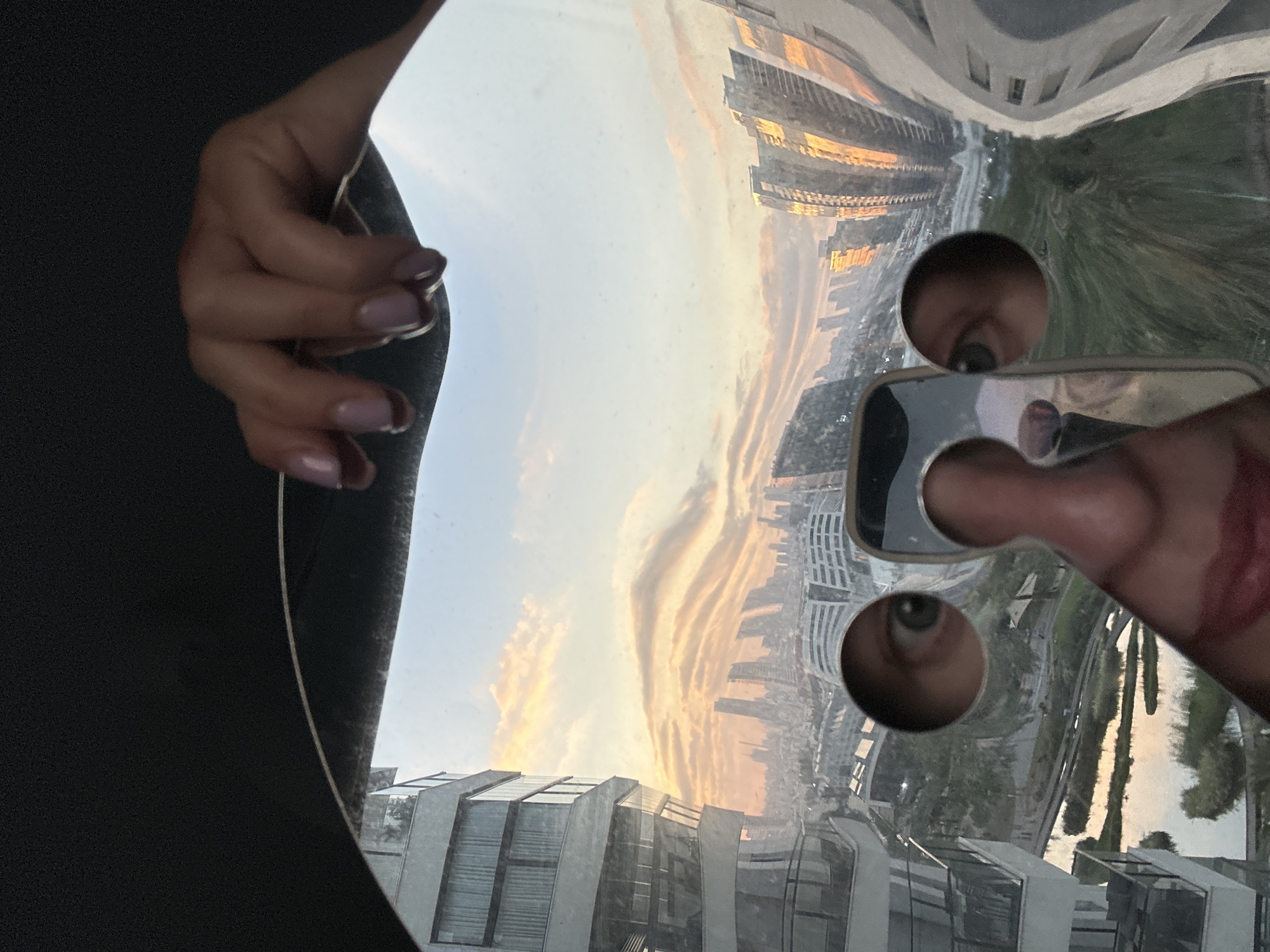 This image is a first-person perspective of a hand holding a smartphone, reflecting a distorted urban scene at sunset. The hand, with light purple-tinted nails, is visible in the upper left quadrant, suggesting the photographer. The smartphone's screen and case are also reflected, showing a distorted view of the photographer's eye and a portion of their lips.
The reflection in the curved, metallic surface reveals a cityscape dominated by modern architecture with glass facades and geometric shapes, indicative of a contemporary urban environment. Tall buildings are visible, some illuminated with warm, golden light, characteristic of sunset. The sky is painted with soft hues of orange, pink, and yellow, forming dramatic, layered clouds. Greenery, likely trees or a park, is also present, adding a natural element to the urban landscape.
The reflection is highly distorted due to the curved surface, creating a fisheye-like effect that warps the buildings and the sky. The time of day is clearly sunset, given the warm lighting and cloud colors. The overall impression is one of artistic self-reflection and an appreciation for urban beauty at a specific time of day. The specific location cues are not strongly identifiable beyond a general urban setting in Petah Tikva, Israel, as provided. There is no visible text in the image.