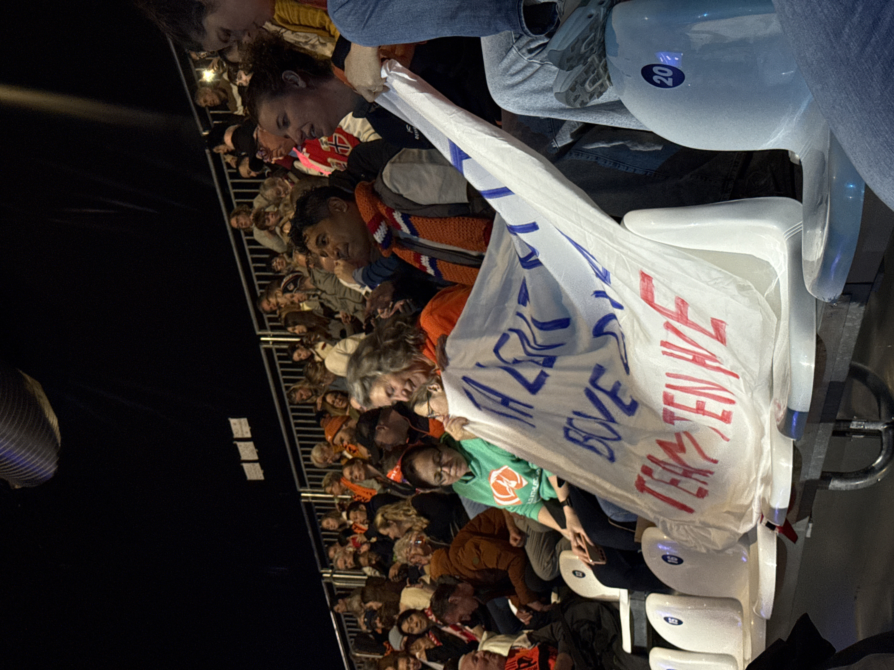 The image captures a section of an indoor stadium or arena in Rho, Italy, during a dimly lit event, featuring a crowd of spectators. Approximately 50-70 individuals are visible, seated in tiered white plastic chairs, some marked with blue numbers (e.g., "20" on a seat in the immediate foreground).
A large white fabric banner, bearing hand-painted blue and red text, is prominently displayed across the mid-ground. Visible blue text includes "TA ZEKR," and visible red text includes "TEAMS TEN HVE."
In the immediate foreground, the lower body of a person wearing light blue jeans and grey athletic shoes is seen, with their right foot resting on a white seat numbered "20." This person appears to be holding the right side of the banner. To their left, a woman with dark, curly hair, wearing a dark top, holds the banner with her left hand, looking towards the left side of the frame. Adjacent to her left, an older woman with grey, wavy hair, dressed in an orange top, smiles while looking towards the banner. Further to the left, a woman with dark hair and glasses, wearing a green hooded sweatshirt with an orange logo, is seated, holding a smartphone with both hands.
To the right of the banner-holding woman, a man with dark hair and complexion, wearing an orange, blue, and white striped knitted scarf over a dark jacket, is observed looking upward and to his right, with his mouth slightly open. Many attendees in the mid-ground and background are wearing orange clothing or accessories, suggesting collective support. The environment is characterized by low ambient lighting, with distinct beams of light from unseen overhead spotlights illuminating sections of the upper arena. Metal railings are visible, separating seating sections in the background.