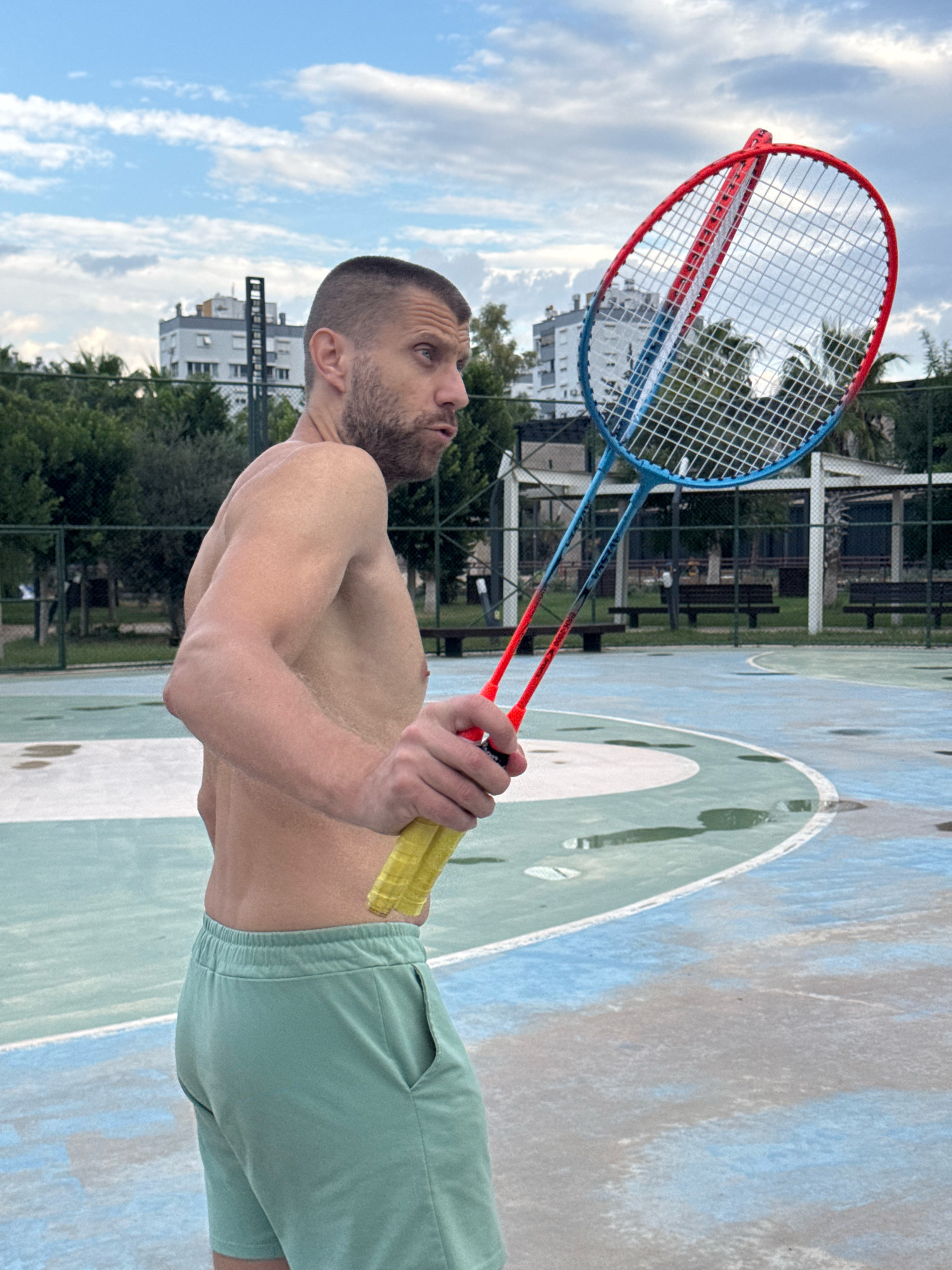 A shirtless man with short hair and a beard stands on a wet outdoor court holding a badminton racket. He is wearing light green shorts. The court has blue and green markings. In the background, there are trees, buildings, and benches. The sky is cloudy. The scene is located in Antalya, Turkey.