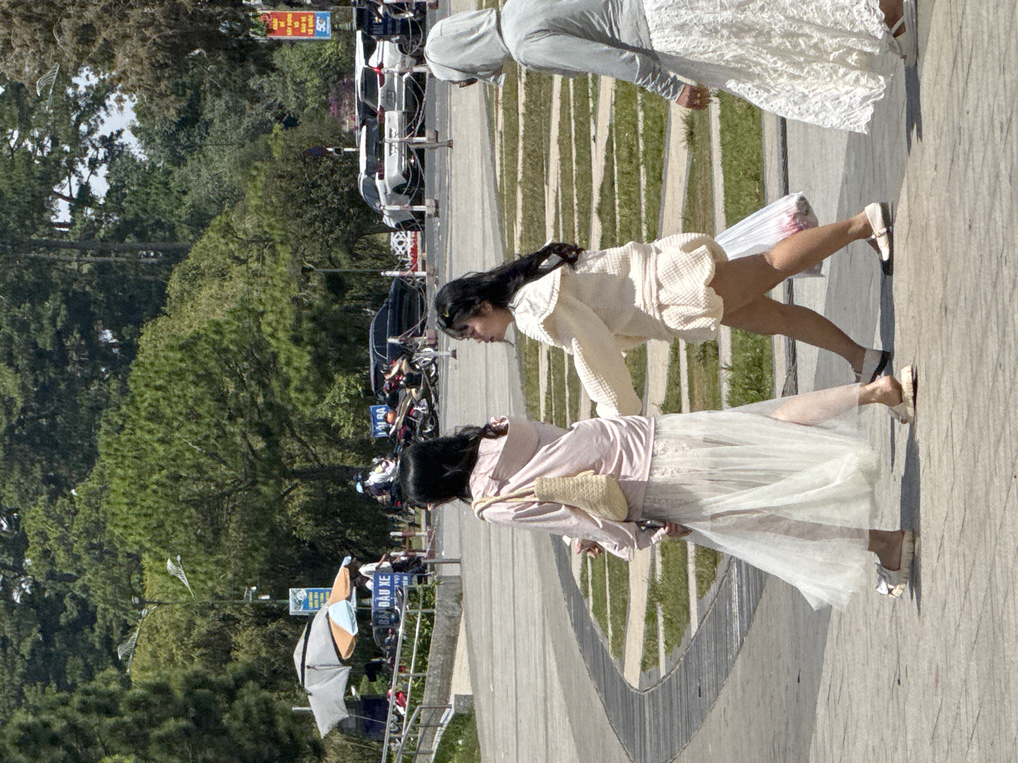 Three women are walking on a paved pathway in Da Lat, Vietnam. The woman on the left wears a long, sheer white skirt and a pink hooded jacket, carrying a straw bag. The middle woman is dressed in a cream-colored outfit, including a ruffled top and shorts, and carries a plastic bag with pink items inside. The woman on the right wears a grey hooded garment and a white lace skirt. In the background, there are trees, parked cars, and motorcycles. A sign with Vietnamese text is visible near the parked vehicles. The pathway has a curved design with grass patches and wooden planks.