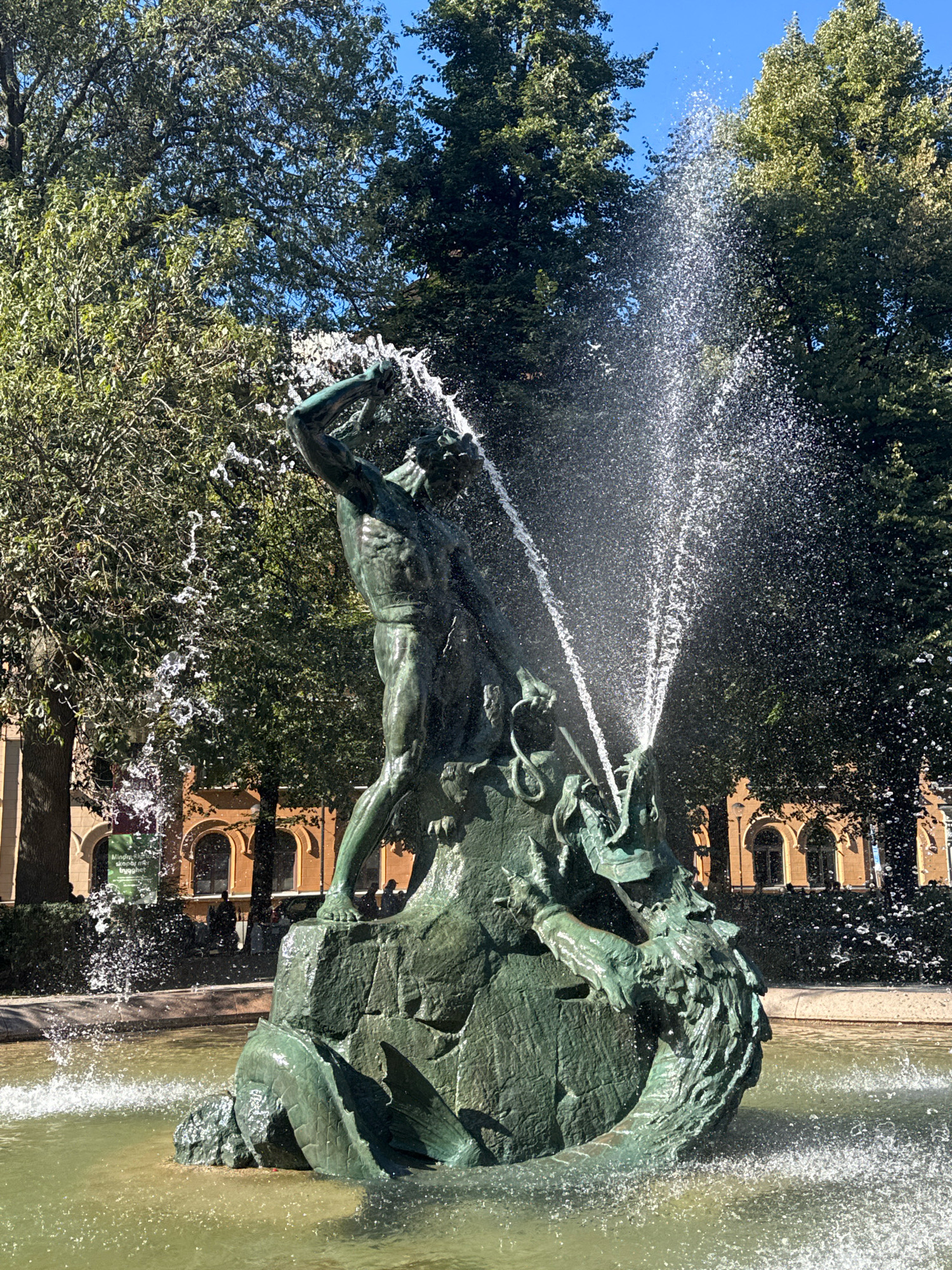 This is a photograph of the Bronsormen fountain, located in Kungsträdgården park in Stockholm, Sweden. The central subject is a bronze sculpture of a muscular man in a dynamic pose, appearing to wrestle with a sea serpent or dragon. The sculpture is depicted as a scene of struggle, with the man holding his arm up, possibly wielding a weapon or bracing himself. Water is actively spraying from the sculpture, creating a fountain effect that obscures parts of the figures and adds a sense of movement and drama to the scene. The sculpture is situated within a pool of water, which reflects the sunlight and the surrounding environment. In the background, lush green trees with dense foliage form a canopy, and the blue sky is visible through the branches. Behind the trees, an architectural structure with arched windows and warm-toned walls is discernible, suggesting a building or facade. Several indistinct figures are visible in the middle ground, standing or walking along a path, adding a sense of scale and human presence to the park setting. The overall scene captures a vibrant and lively moment in a public urban space, highlighting the artistic and natural elements of the location.