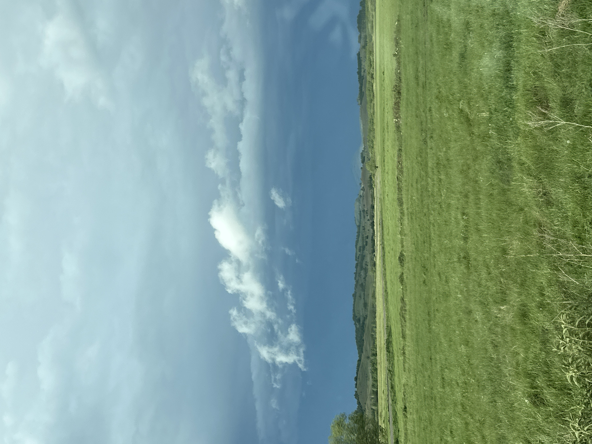The image shows a landscape with a clear sky with some white clouds covering about half of the sky and a green grass-covered field. The ground is to the right of the image and slopes down to the left, suggesting a hill or slight elevation. The sky takes up a large portion of the picture with gradients of white to blue. The location is unknown. No specific details about cities or people are identifiable.
