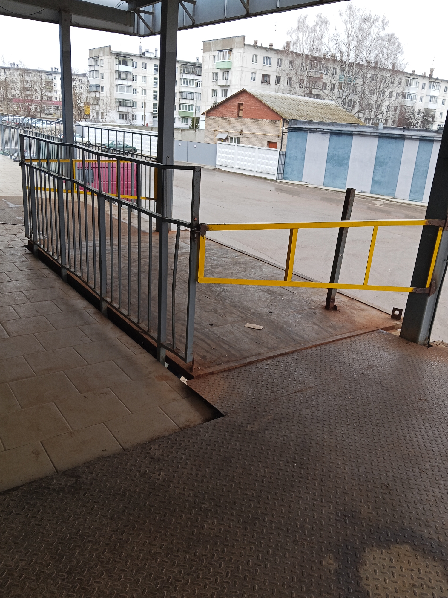 The image shows a metal ramp with railings leading up to a higher platform, likely for loading or unloading goods. The ramp and platform are made of metal with a diamond plate pattern. The railings are gray with yellow accents on one side. In the background, there are apartment buildings and a wall with blue and white stripes. The location is an urban area. No people are visible.