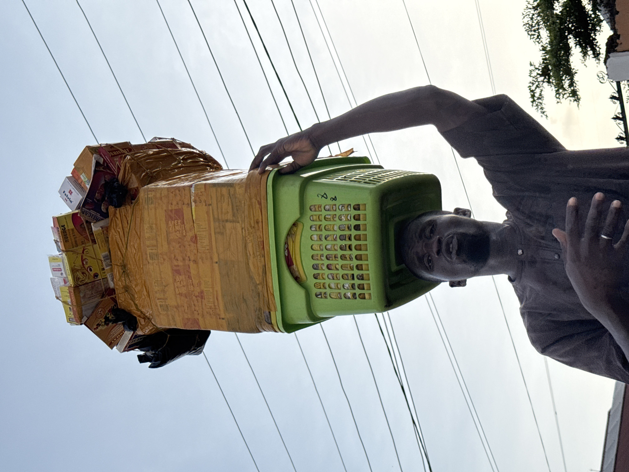 A man in Ado, Nigeria, is carrying a large bundle of goods on his head. The bundle is balanced on a green plastic basket. The goods include boxes and items wrapped in yellow tape and plastic. The man is holding the basket with one hand. He is wearing a brown shirt. Overhead power lines are visible against the sky.