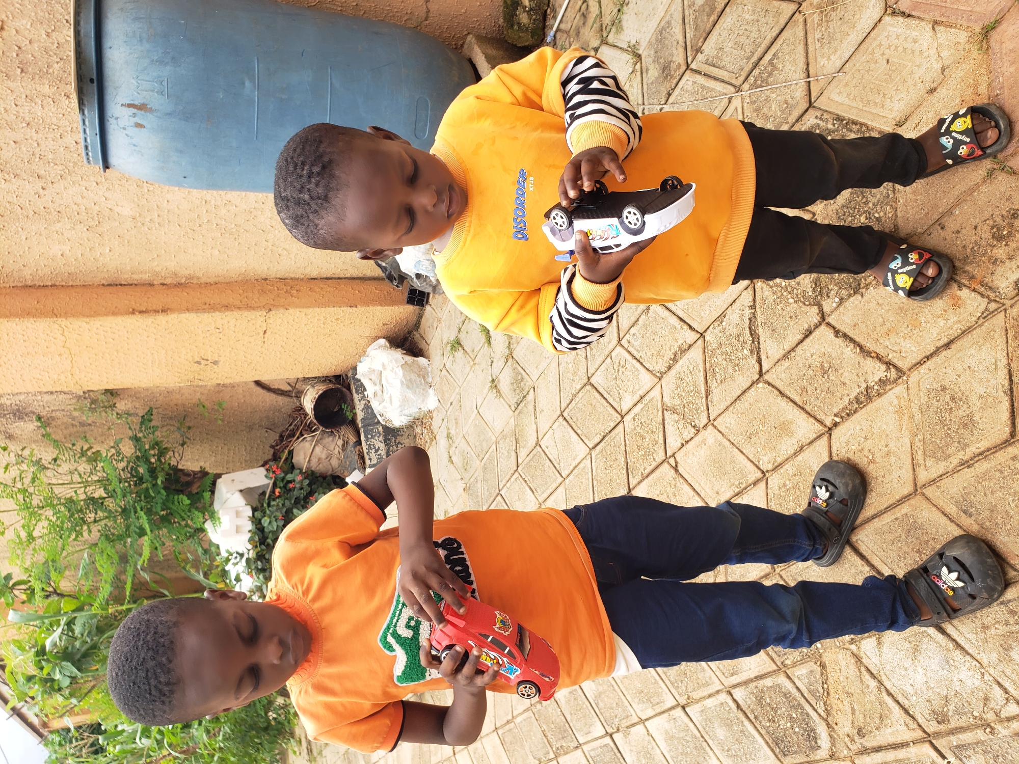 Two young African boys are standing on a tiled walkway outside. The boy on the left, wearing an orange t-shirt and blue jeans, is holding a red toy car. He is looking down at the car. The boy on the right, wearing a yellow sweatshirt with black and white striped sleeves and black pants, is holding a black and white toy car. He is also looking down at the car. Behind the boys, there is a textured yellow wall, some greenery, and a large blue barrel. The ground is paved with square tiles. This scene is likely in Sabon Tasha, Nigeria, given the context.