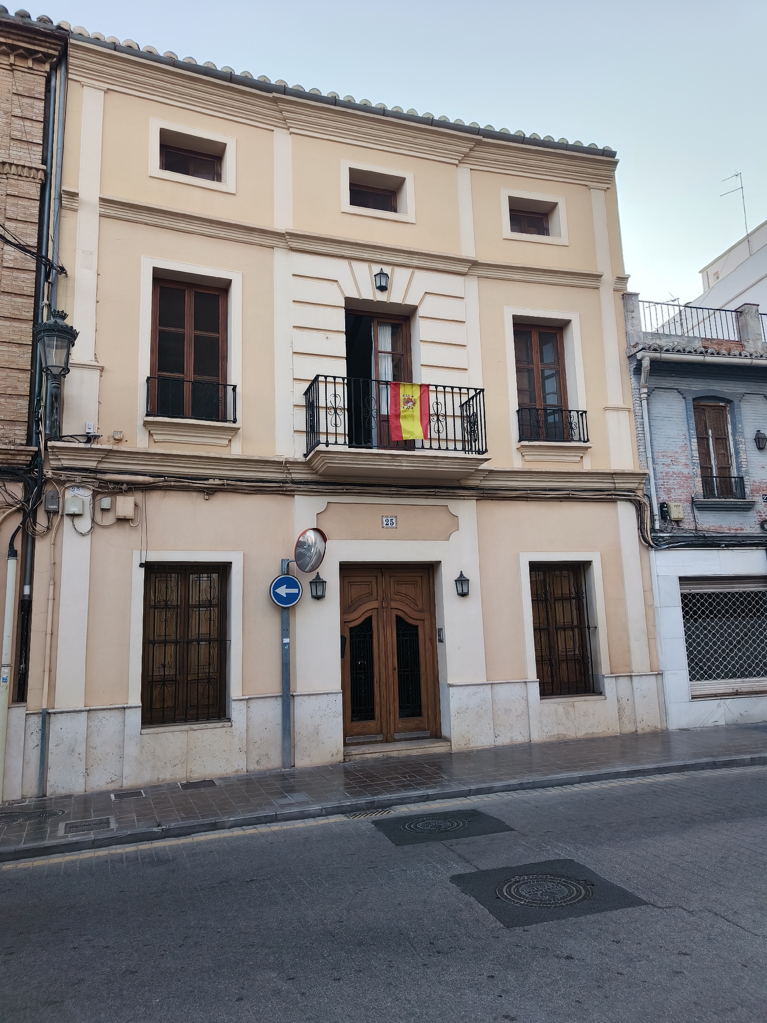 The image shows the facade of a two-story building in Benimàmet-beniferri, Spain. The building is painted in a light yellow and cream color. The lower story has a large wooden double door in the center, flanked by two windows with metal grates. Above the windows and doors, there is a decorative cornice. The upper story has three windows. The central window is an open balcony with a Spanish flag hanging from it. There are also two smaller windows on either side of the central balcony. The building is situated on a street with a tiled sidewalk and asphalt road. A street lamp and a directional sign are visible on the sidewalk in front of the building. The sky is a pale blue.