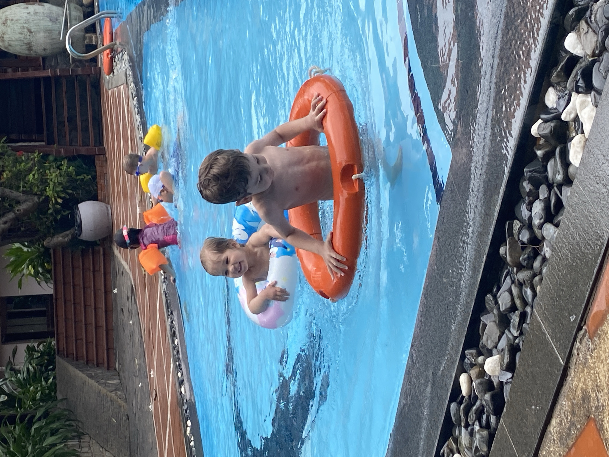 Two young, shirtless boys are swimming in a pool, located in Phu Thuy Ward, Vietnam. The older boy has short brown hair and is holding onto an orange life ring. The younger boy is wearing a blue and white inner tube. The pool is bordered by dark grey tiles and black and white pebbles. In the background, other children are in the pool. Vegetation and a building constructed of orange brick can be seen in the background. The water is light blue.