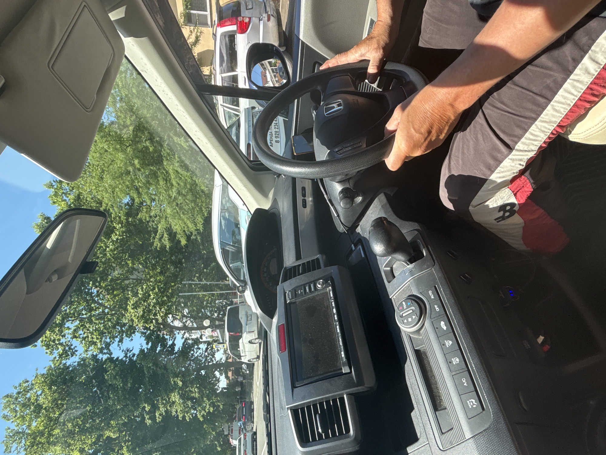 The image shows the interior of a car, likely a Honda, with a person's hands on the steering wheel. The driver is wearing a dark shirt with red and white stripes on the sleeves. The steering wheel is black with a Honda emblem in the center. A car's rearview mirror is visible, reflecting trees and buildings.
In the foreground, a car's dashboard is visible, featuring a touchscreen infotainment system and climate controls. The climate controls have buttons labeled "AUTO," "OFF," "TEMP," "A/C," and what appears to be a fan speed dial. Visible text on the dashboard includes "AUTO" and "A/C". Through the windshield, other cars and trees are visible, suggesting the car is on a street or in traffic. The lighting indicates it is daytime, likely sunny, given the shadows and brightness. The presence of trees and the general environment suggest an outdoor setting, possibly in a city.
The context provided places the scene in Sochi, Russia. The image captures a moment from the driver's perspective, offering a glimpse into the car's interior and the external environment. There is no clear indication of a specific event or strong emotional cues, beyond the practical act of driving.