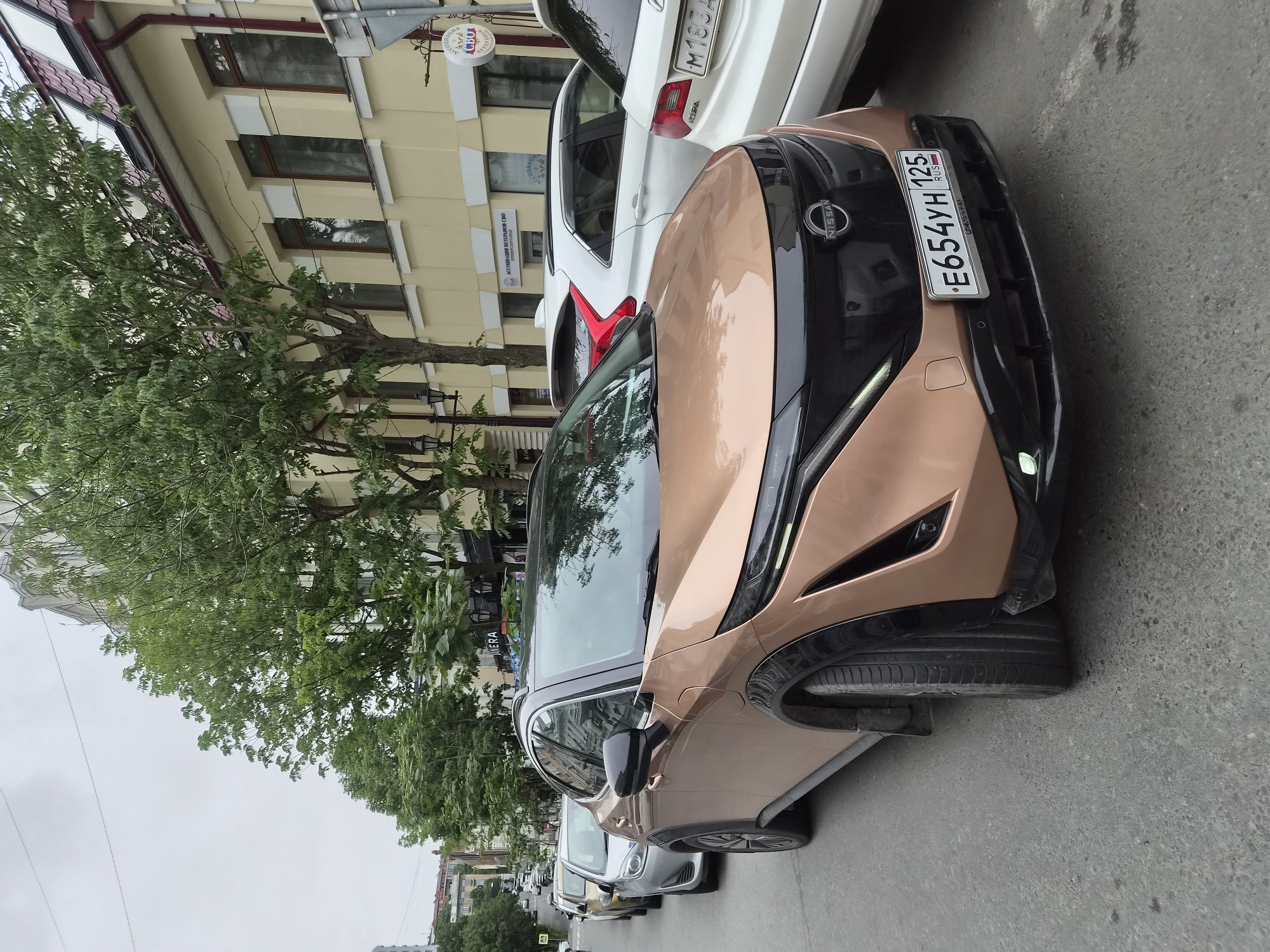 A group of cars are parked in a line on a city street in Vladivostok, Russia. The foreground features a copper-colored Nissan Ariya with a license plate that reads "E654YH 125". Behind it, a silver sedan and a white SUV are visible. The street is paved and lined with trees and buildings. The buildings appear to be a mix of historical and modern architecture, with one building in the background featuring a yellow facade and red-tiled roof. The sky is overcast. There are no people visible in the image. The overall scene depicts a typical urban streetscape.