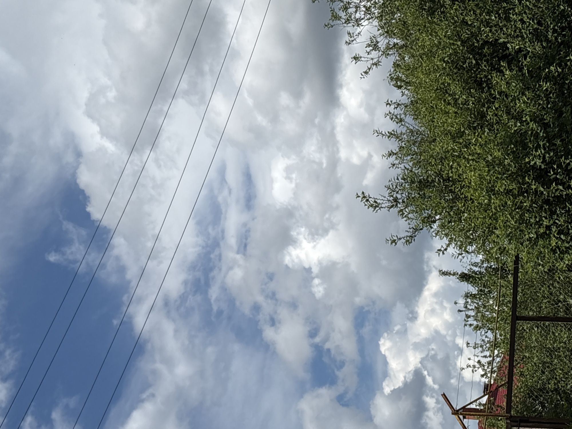 The image shows a view looking upwards toward the sky in Прикамский, Russia. Power lines run diagonally across the left side of the frame. The sky is partly cloudy, with areas of both blue sky and puffy white clouds. On the right side of the frame, a tree with dense green foliage is visible. At the lower right, a rusty metal fence and a partially visible red roof suggest the presence of a building.