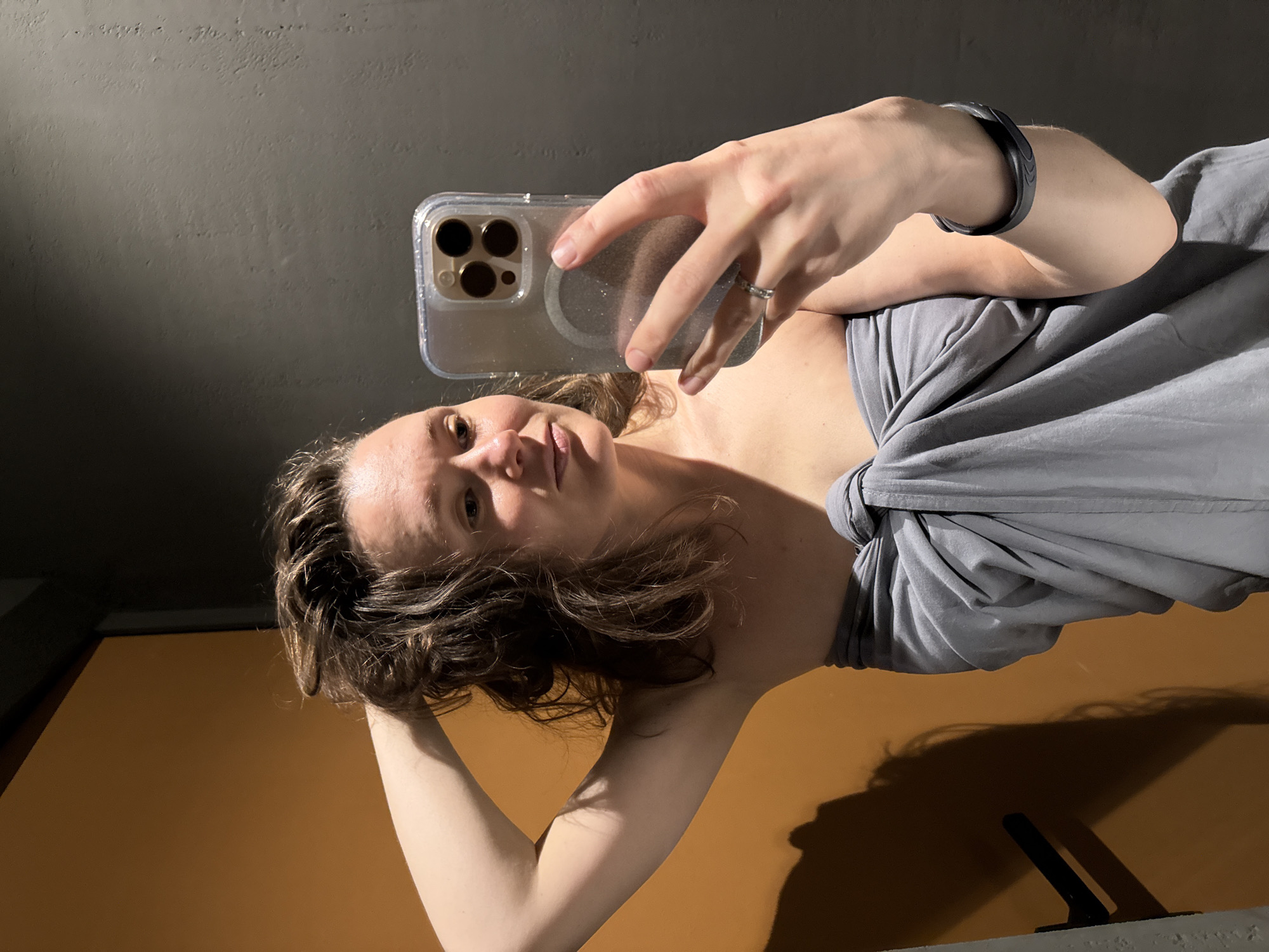 A woman is taking a selfie with a smartphone in a clear case that reveals three camera lenses. She is wearing a grey top with a knot at the shoulder, and a black bracelet on her left wrist. The background is a dark grey wall on top and a brown wall on the bottom. Her hair is short and brown. Her left hand is holding the back of her head. There is a ring on her finger. The image does not contain sufficient information to determine the location as Vyakhtelevo, Russia, or to provide a detailed description of actions or interactions.