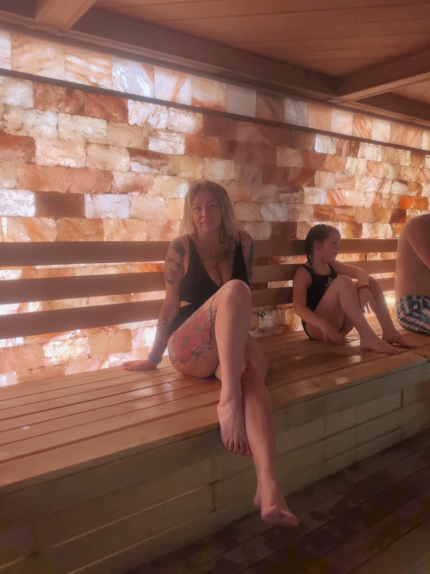 The image depicts three individuals inside a halotherapy room, indicated by the wall constructed from what appears to be salt bricks. A woman is seated on a wooden bench, directly facing the camera with legs crossed and tattoos visible on her arms and leg. To her right, a young girl is also seated, looking to her right. The right side of a man is partially visible on the right edge of the scene. The room has a wooden ceiling and the floor is wooden. The location is unknown.