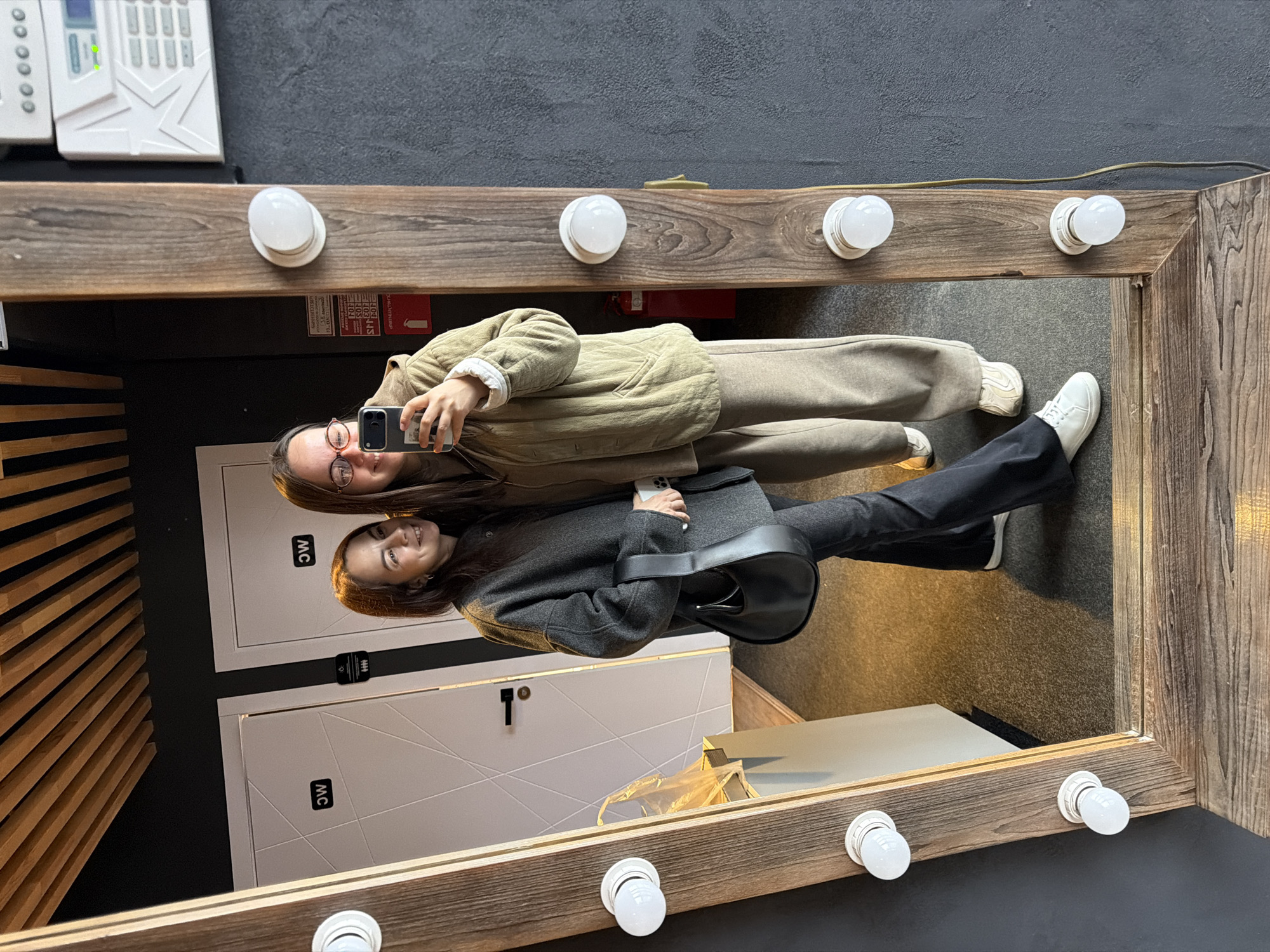 Two women are reflected in a mirror with lights around the frame. One woman is wearing glasses, a tan jacket, and pants, and is holding a phone. The other woman is wearing a gray coat, black pants, and a black purse. The background shows restroom doors with "WC" signs. The location is Chaikovsky, Russia.