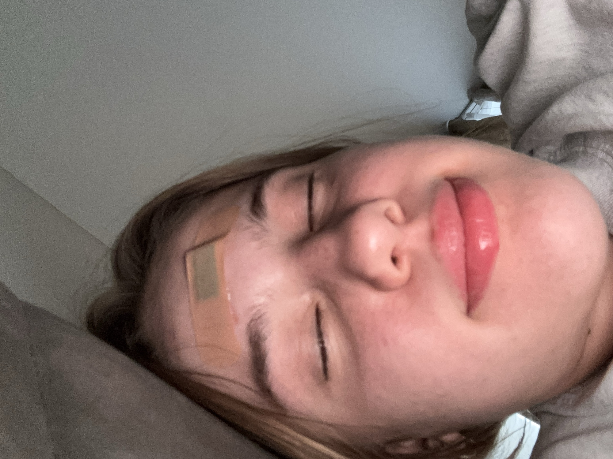 The image is a close-up selfie of a young person with their eyes closed, lying down, with a light-colored blanket or pillow over their head and shoulders. A band-aid is visible on their forehead. The person has blonde hair, fair skin, and is wearing a grey hooded sweatshirt. The background is out of focus and appears to be a light-colored wall or ceiling. There is no indication of the image being taken in Lednevo, Russia.