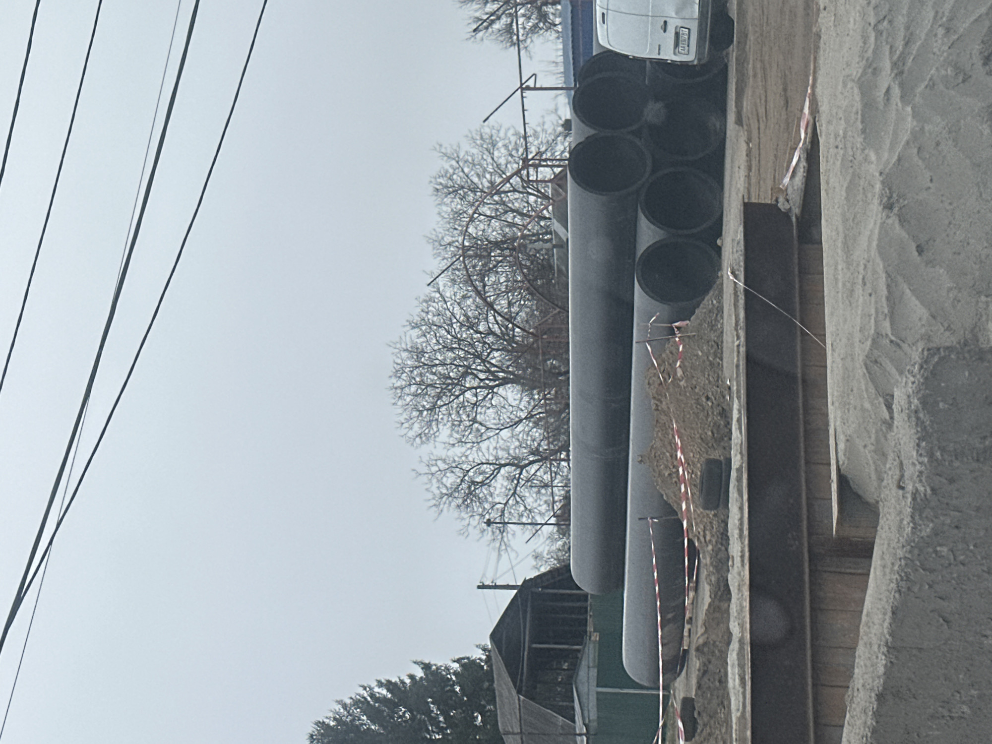 The image shows a construction site with large black pipes stacked alongside an excavated trench reinforced with steel beams. Red and white striped caution tape borders the site. A white van is parked nearby. Overhead, bare tree branches reach towards a grey sky, with power lines visible. The setting appears to be an urban environment, with a building partially visible behind the pipes. A license plate on the white van is visible, with the number AA 0627 BI. Given the license plate, this image is likely taken in Kyiv, Ukraine.
