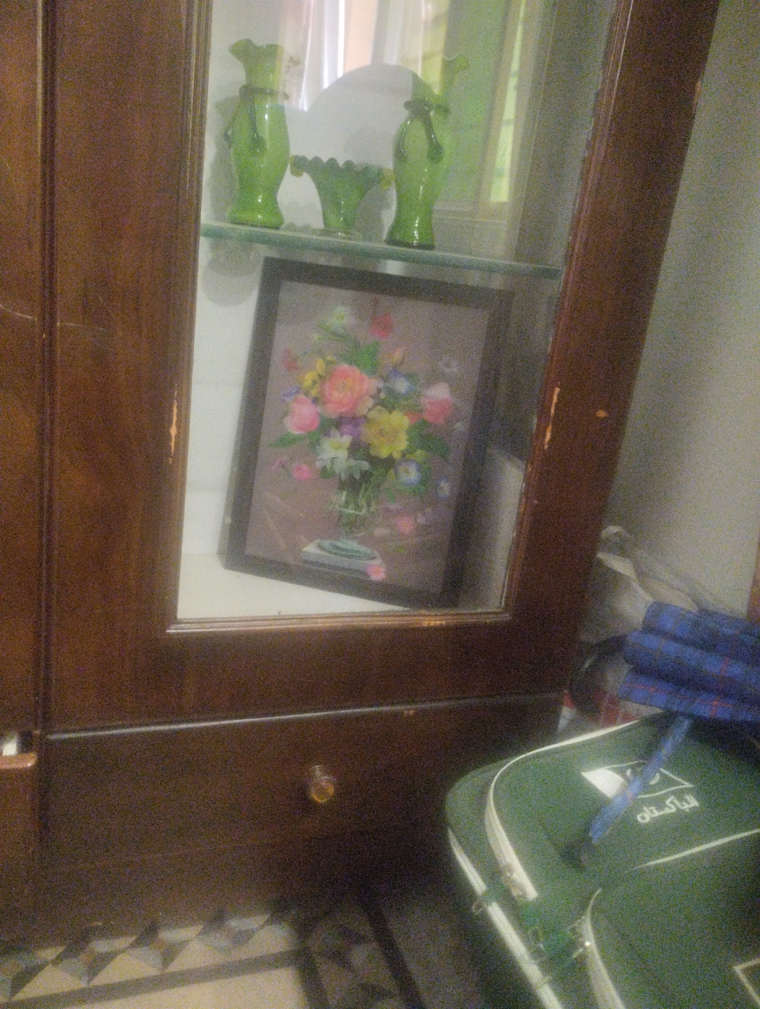 A framed floral painting is displayed on a glass shelf inside a wooden cabinet. The painting depicts a vase filled with various colorful flowers, including pink, yellow, and orange blossoms, against a muted background. To the left of the painting, on the same shelf, are two green glass vases. The left vase is taller and has a rounded shape with a decorative element around its neck. The right vase is shorter with a ruffled opening. The cabinet is made of dark wood with visible grain. In the foreground, a green and white bag with the word "پاکستان" (Pakistan) printed on it is partially visible. The floor has a patterned tile design. The lighting in the scene is somewhat dim, creating reflections on the glass surfaces. The overall impression is of an indoor display cabinet.