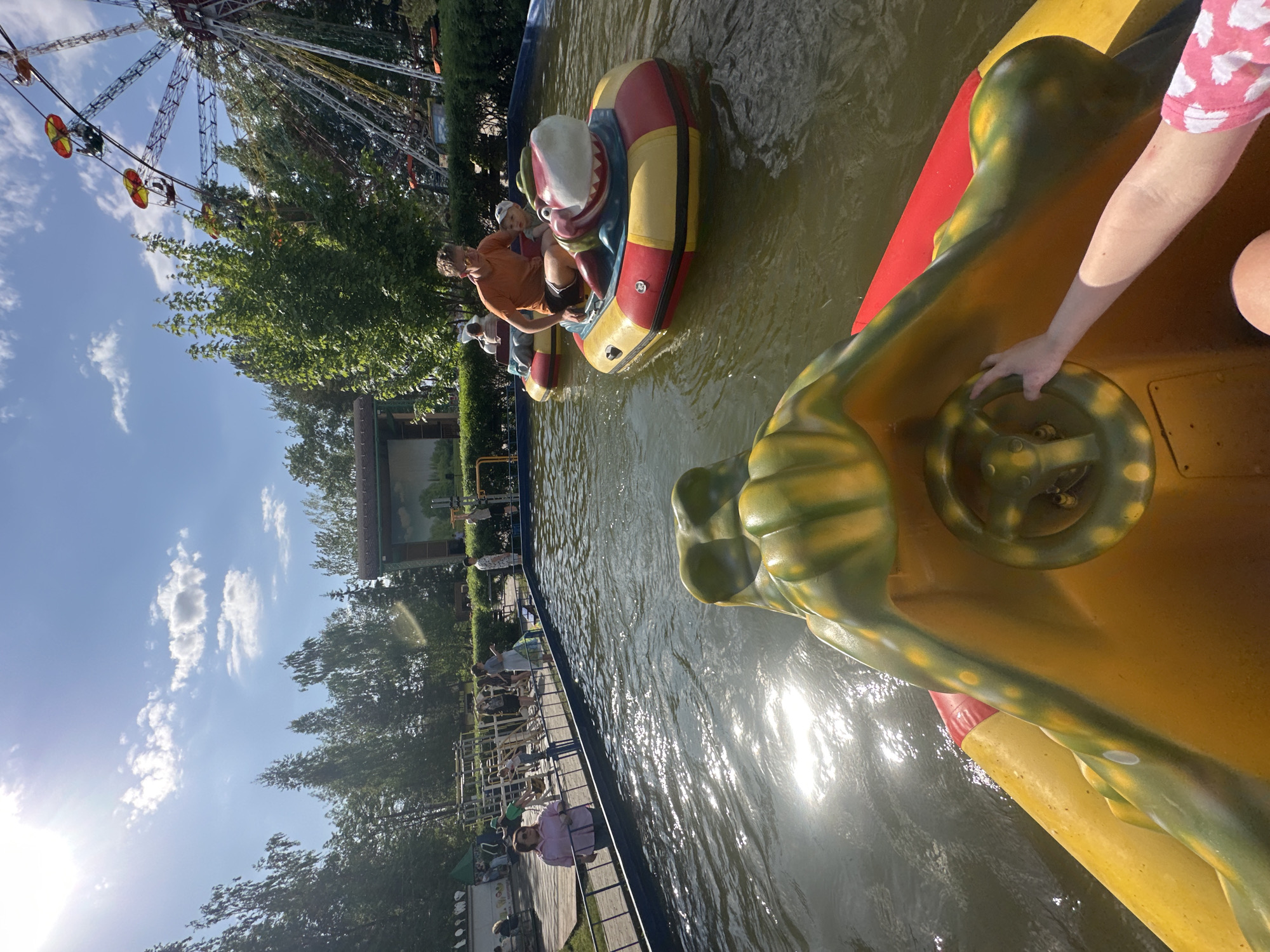 A high-angle shot depicts a water ride with themed boats at what appears to be an amusement park, potentially located in Chaikovsky, Russia. In the foreground, a yellow and green boat with a steering wheel is occupied by a person whose arm and partial torso are visible. Their hand is on the steering wheel. The boat is brightly colored with a design that suggests a frog or similar creature. The water is murky, with light reflecting off the surface. Further away, other boats are occupied by people. One boat has an adult male in an orange shirt and a child in it. In the background, a Ferris wheel, trees, and buildings are visible. A line of people can be seen alongside a railing beside the water. The sky is blue with scattered clouds, suggesting a sunny day.