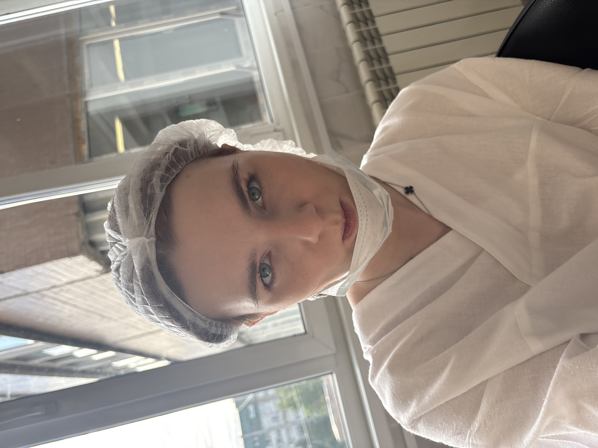 An eye-level portrait shows a person, likely female, indoors, looking directly at the camera with a neutral expression. The image is significantly tilted to the left. The individual has fair skin, light-colored eyes, and visible eyebrows. They are wearing a white disposable bouffant cap covering their hair, a white disposable medical gown or smock, and a white medical face mask pulled down to rest below their chin, revealing their nose and lips. A delicate silver chain with a black four-leaf clover pendant is visible around their neck.
The setting appears to be a clinical or cosmetic environment, possibly in Saint Petersburg, Russia. Behind the person is a large window with multiple panes, through which an urban outdoor scene is visible, including hints of other buildings and foliage, suggesting a daytime setting. Reflections in the window show parts of the interior, including what might be a textured or tiled white wall and some ceiling elements or light fixtures. The person appears to be seated, with a dark, possibly padded, surface visible behind their right shoulder, which could be part of a chair or headrest. The bright, natural light from the window illuminates the scene. No visible text is present in the image.