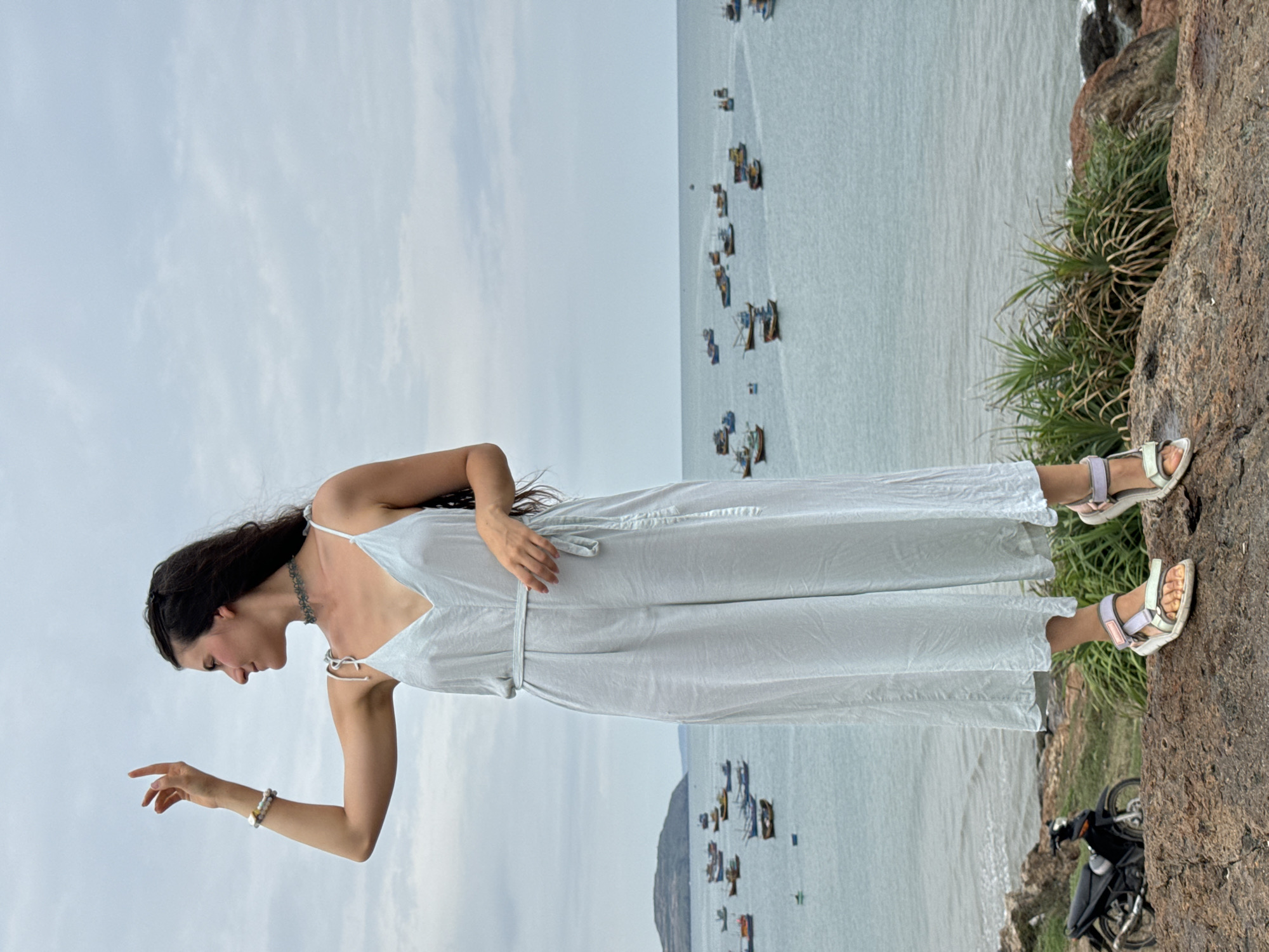A woman with dark hair and a white jumpsuit stands on a rocky outcrop overlooking the sea. Her right arm is extended with her fingers gently pointed downwards. The ocean is visible behind her, dotted with numerous boats. A motorcycle is parked on the rocks behind her feet. The location is Mui Ne, Vietnam, implied by the presence of the fishing boats typical of the area.
