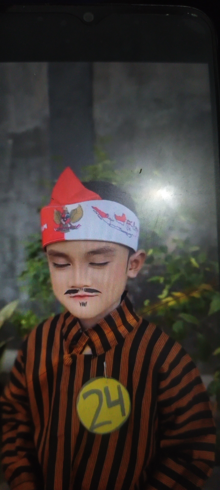 The image shows a young child dressed in traditional Indonesian attire. The child is wearing a striped shirt and a headband adorned with the Indonesian flag and the national symbol, Garuda Pancasila. The child also has a drawn-on mustache and goatee, and a yellow circular badge with the number "24" is attached to the shirt. The child's eyes are closed, and they appear to be posing for a photograph. The background is blurred and suggests an outdoor setting with foliage. The image appears to be taken within the city of Ambulu, Indonesia, likely during a cultural event or celebration.
