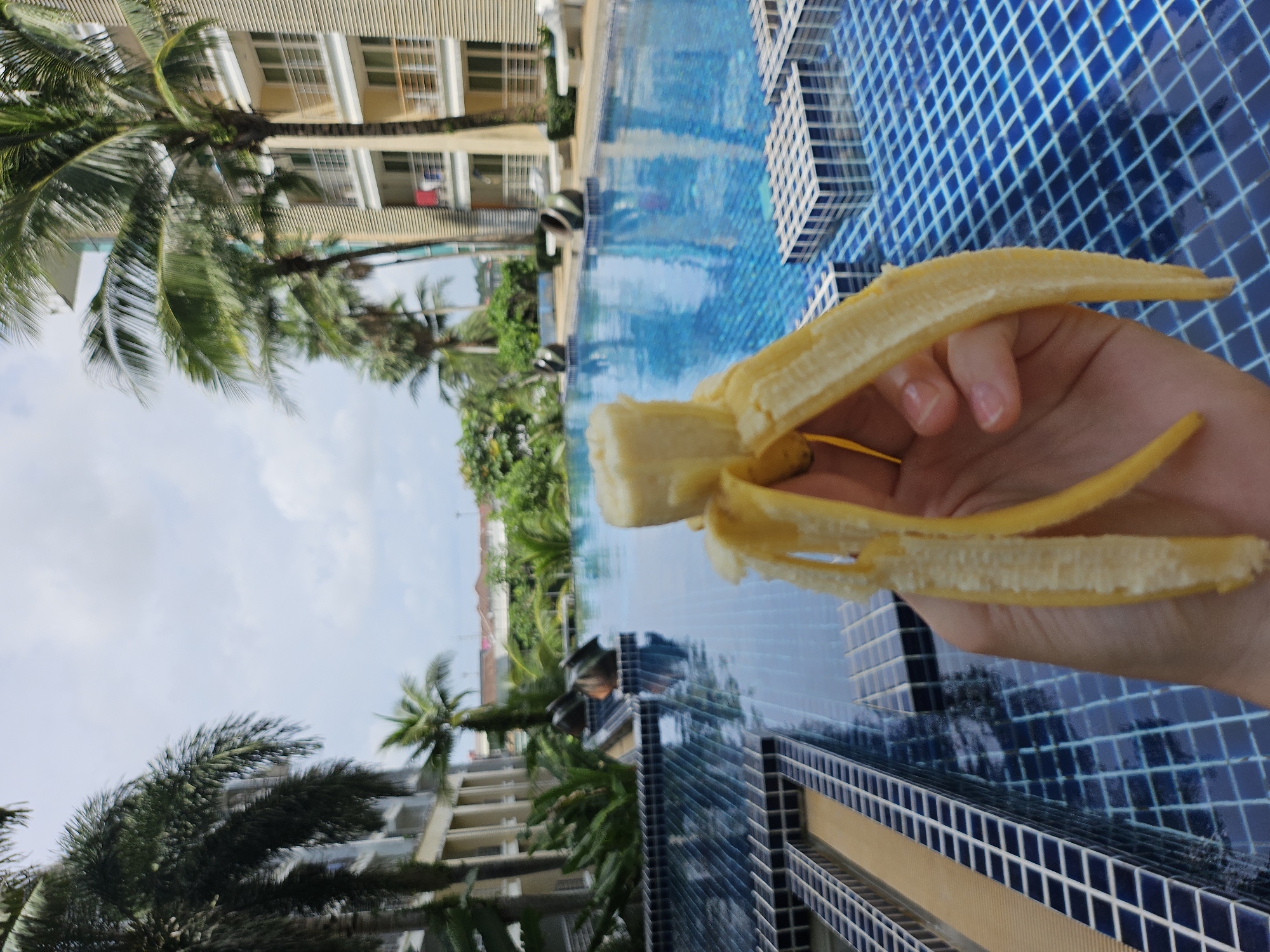 An extreme close-up shot focuses on a partially peeled banana held by a person's hand in the foreground. The banana is yellow and appears soft. In the background, a bright blue swimming pool with a tiled edge stretches out, reflecting the sky and surrounding palm trees. Buildings with balconies and windows, characteristic of a resort or hotel, are visible behind the pool, along with lush green tropical foliage. The scene suggests a location in a warm climate, likely a tourist destination such as Kathu, Thailand, known for its resorts and pools.