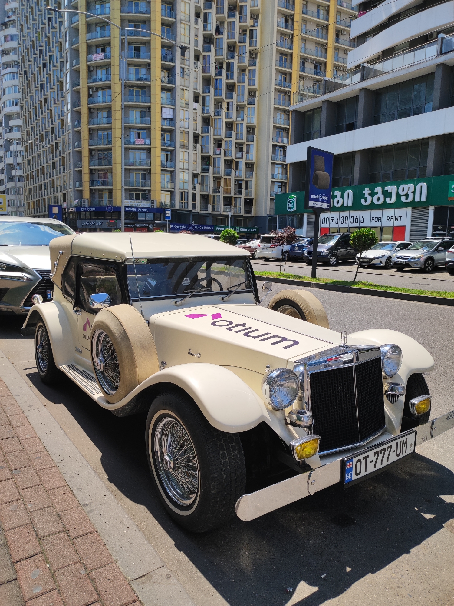 An eye-level, medium shot captures a vintage cream-colored convertible parked on a city street in Batumi, Georgia. The car, with its chrome accents and wire-spoke wheels, is the central subject, its ornate design contrasting with the modern architecture in the background. The building behind the car features rows of balconies, and signs for businesses are visible, including "QoQoU Bakery" and "FOR RENT". A street lamp stands to the left of the frame, and other cars are parked in the background. The car's license plate reads "OT-777-UM". The ground is paved with red brick on the left and asphalt on the right, with a concrete curb separating the two. The scene is illuminated by bright daylight, casting shadows on the street. No people are visible in the frame.