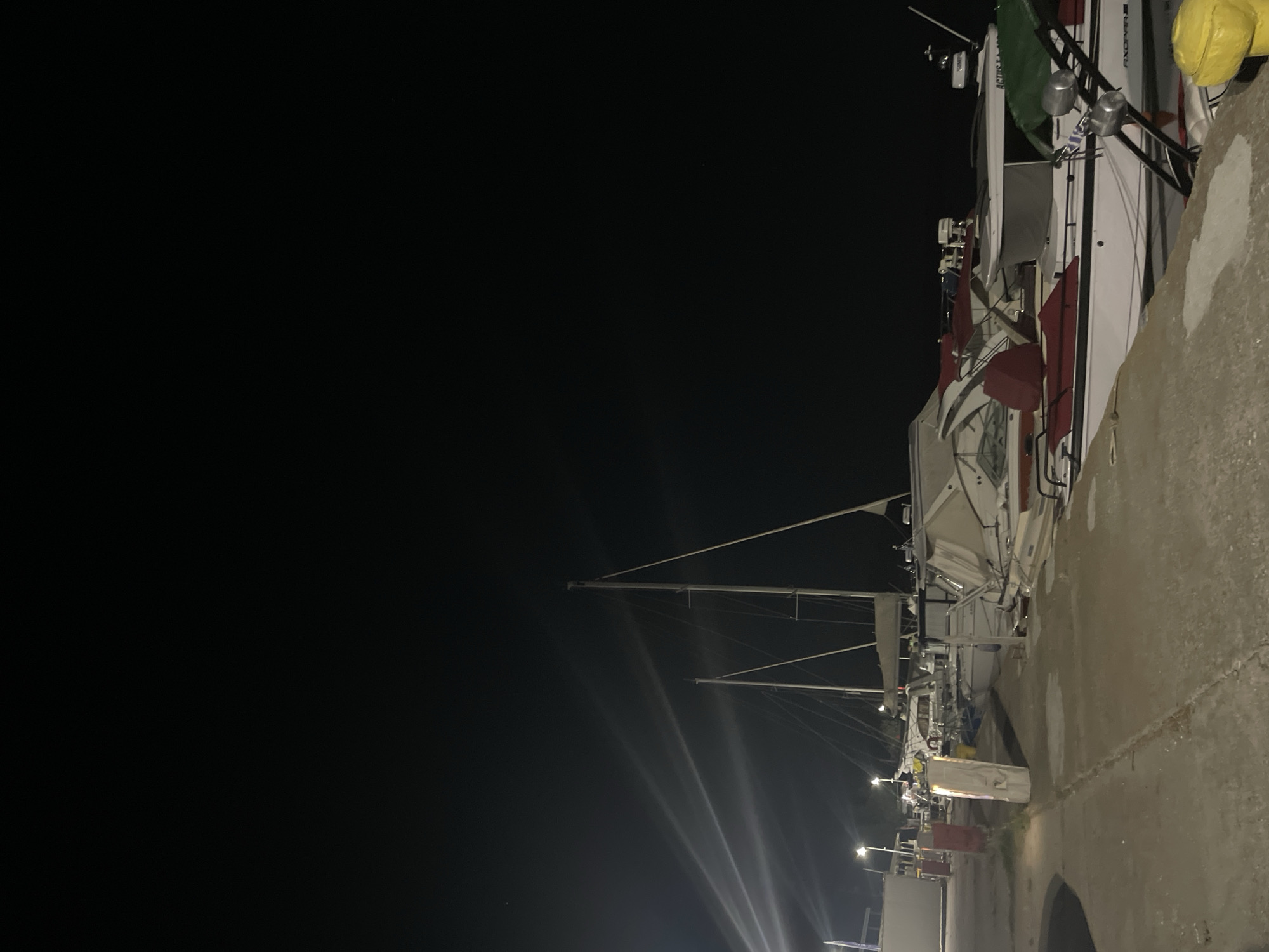 In Limenas Avdiron, Greece, at night, several boats are docked in a harbor. The scene is illuminated by artificial lights. In the foreground, a concrete walkway runs along the edge of the harbor. A yellow bollard is visible on the right side of the walkway. Several boats are moored alongside the walkway, including a white motorboat with red seating and a green cover. Further back, two sailboats with tall masts are visible. The sky is dark, with some light beams from unseen sources cutting through the darkness. The overall impression is of a quiet harbor at night.