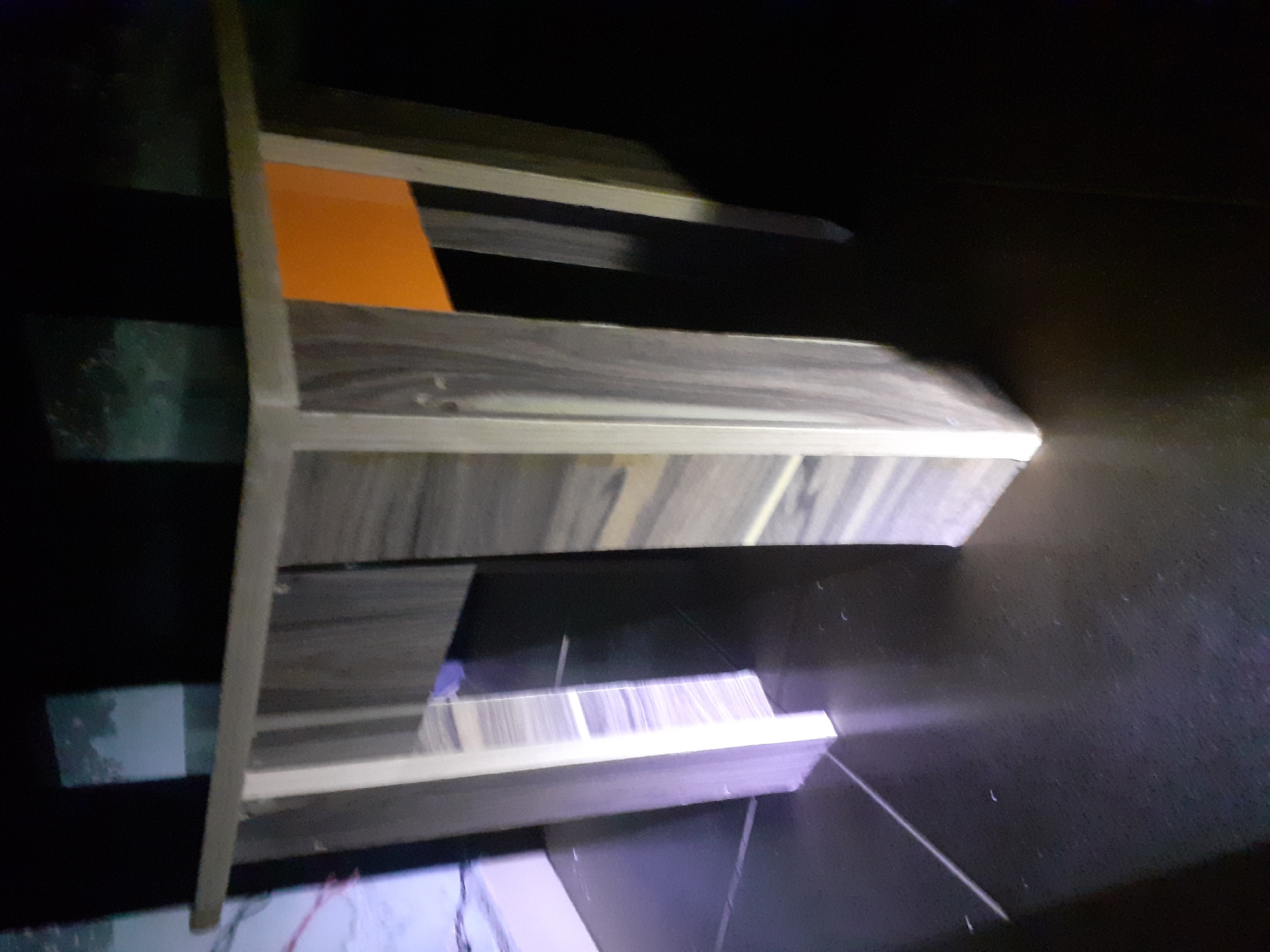 The image shows a corner of a dark room, focusing on a three-legged table. The table has a wooden top with a grain pattern and a lighter-colored edge. The legs are also made of wood, with visible grain patterns on some and plain lighter wood on others. One side of the table has an orange panel. The floor is dark and reflective. In the background, there are dark, indistinct shapes, possibly furniture or walls, with a hint of what appears to be a patterned surface on the left side of the frame. The lighting is dim, with light sources illuminating the table legs and parts of the tabletop. There are no people or animals visible in the image.