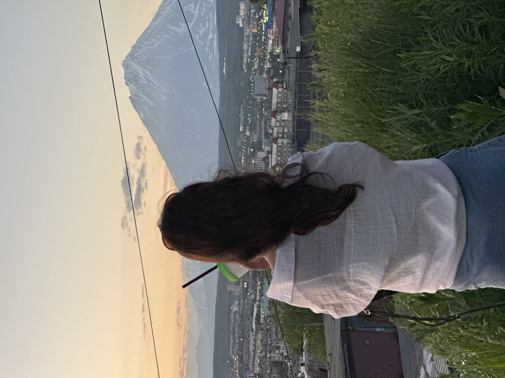 The image shows a woman from behind, facing a panoramic view of Petropavlovsk-Kamchatsky, Russia, at sunset. She is holding a cup with a straw to her lips. The woman has long, dark hair and is wearing a white off-the-shoulder shirt and denim shorts. In the background, the city lights are twinkling, and a snow-capped volcano, likely one of the Avachinsky volcanoes, dominates the skyline under a soft, orange and yellow sky. The foreground is filled with tall green grass, and to the left, there are some garages. Power lines are visible across the sky.