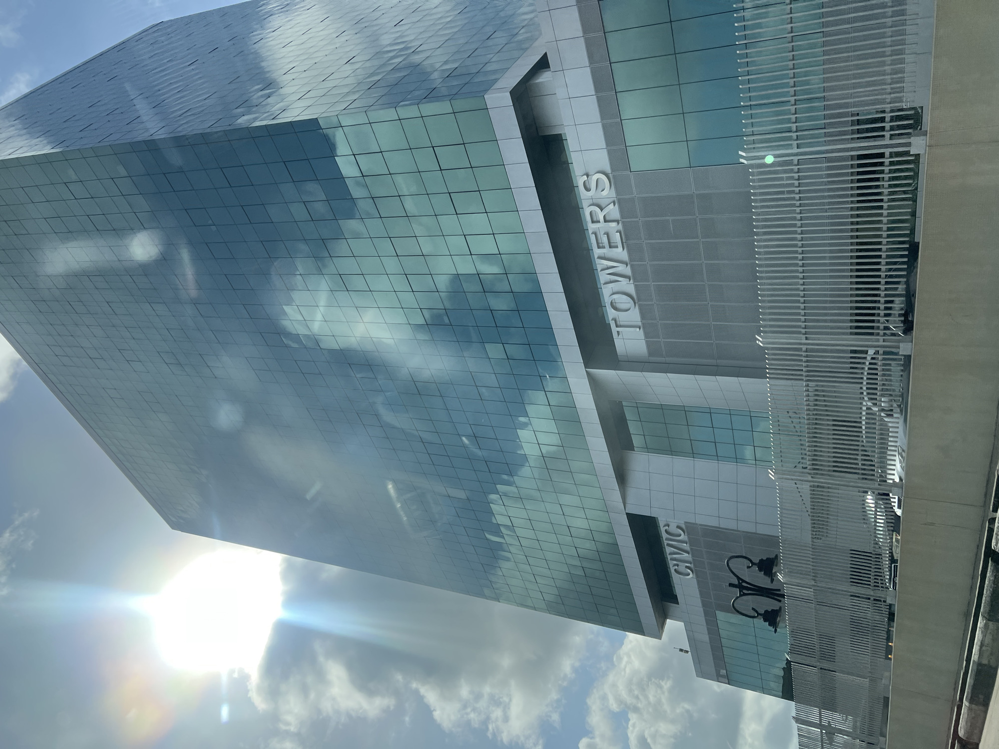 A low-angle view of a building named "Civic Towers," located in Ikoyi, Nigeria. The building has a glass facade with a grid-like pattern, reflecting the sky with clouds. The "TOWERS" and "CIVIC" names are vertically positioned on the side of the building. A white metal fence is visible on the right. Sunlight is visible, creating a bright flare in the lower left corner.