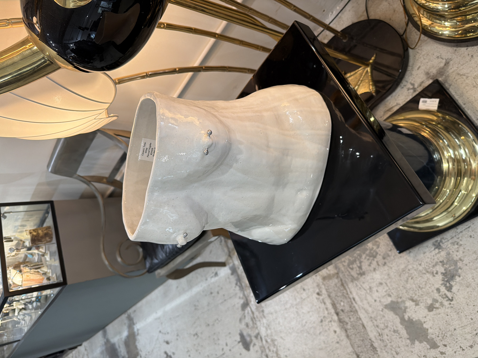 The image shows a white ceramic torso sculpture with pierced nipples displayed on a black square base. It is located indoors, likely in a shop or gallery, as indicated by other decorative objects in the background, including a floor lamp with brass elements and a glass display case. The location is in Los Angeles, United States of America, as indicated by metadata associated with the image. The floor is concrete.