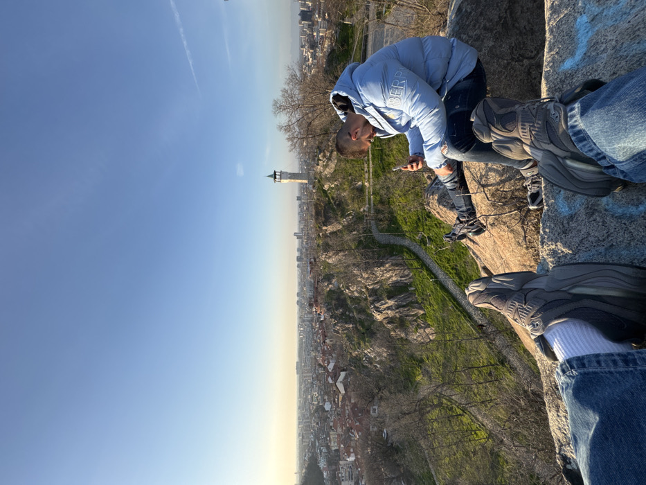 The image displays an elevated panoramic view of the city of Plovdiv, Bulgaria, observed from a rocky outcrop on one of its hills, identifiable by the prominent Clock Tower (Sahat Tepe) in the mid-ground. The perspective is first-person, featuring the lower legs and feet of the photographer, clad in blue jeans, white socks, and dark grey athletic shoes, resting on a foreground rock partially marked with light blue graffiti. To the right, a male subject is seated on an adjacent rock, wearing a light blue puffer jacket with a "BERP" logo on the left chest, ripped dark blue jeans, and dark athletic shoes. He is actively engaged with a white smartphone, holding it with both hands and looking down at its screen. Below the subjects, a steep, green, rocky hillside dotted with bare trees descends, featuring a winding paved pathway. The urban landscape of Plovdiv extends into the background under a clear blue sky, exhibiting a mix of residential and taller structures. The sun appears low on the left horizon, casting a warm glow across the scene, indicating late afternoon. A faint contrail is visible in the upper right quadrant of the sky.