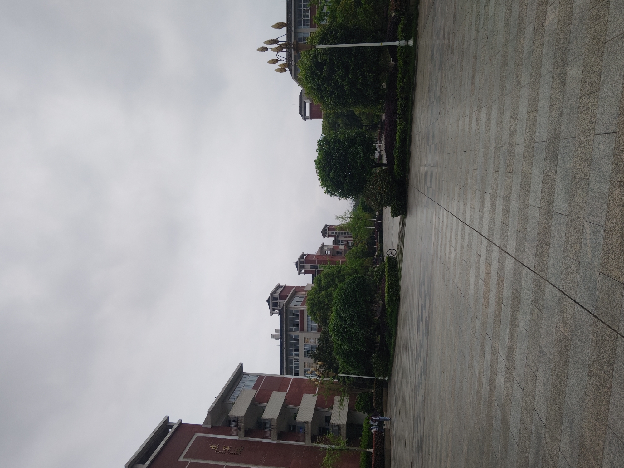 The image depicts an outdoor scene in Chongqing High-tech Zone, China. A wide, paved pathway dominates the foreground, stretching into the distance with tile-like pattern. Buildings constructed primarily of red brick are visible along the pathway. There are some green trees and bushes along the pathway. The sky is overcast. There is at least one bicycle on the pathway. There are also some people standing in front of a building.