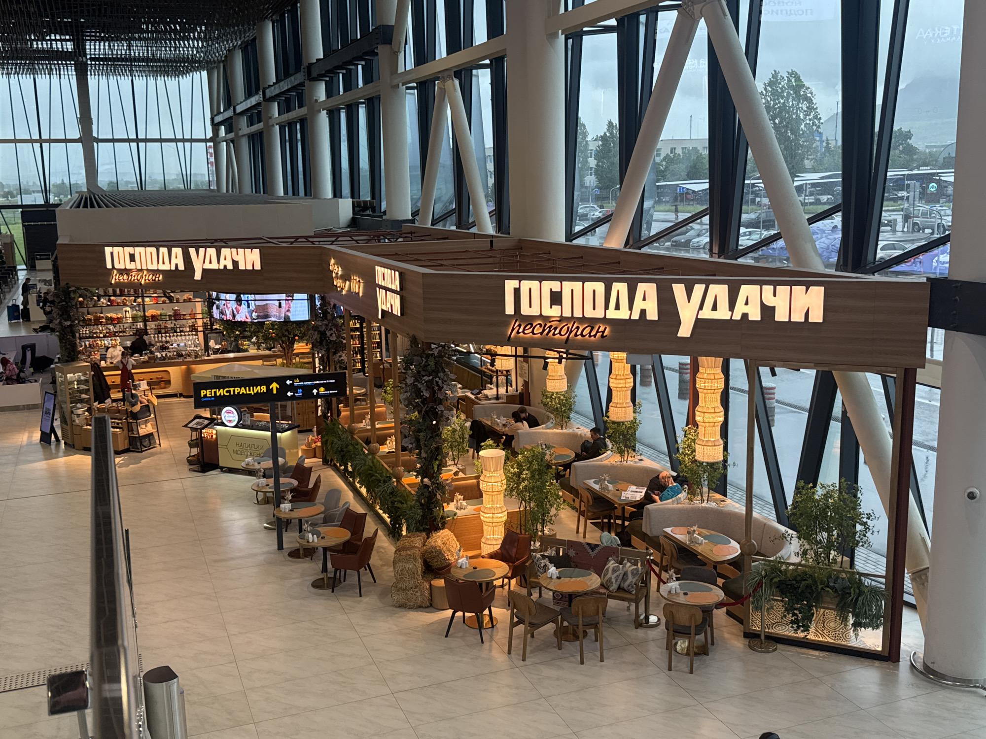 Here is a detailed description of the image:
**Scene:** The image captures the interior of what appears to be a modern airport terminal. A restaurant with a distinct aesthetic dominates the central view.
**Restaurant:** The restaurant is called "ГОСПОДА УДАЧИ" (Gospoda Udachi), which seems to be written in Russian and likely translates to "Lords of Fortune." The decor is a blend of rustic and contemporary, featuring a wooden facade with illuminated signage. Inside, there are tables and chairs arranged in a semi-open layout, with potted plants and decorative lighting adding to the ambiance.
**People:** Several individuals are present within the restaurant, mostly seated at tables. One appears to be reading a newspaper. There are other people visible in the background, likely airport staff or passengers.
**Objects:** The scene contains a variety of objects, including tables, chairs, sofas, decorative plants, lamps, and a direction sign that includes "РЕГИСТРАЦИЯ" ("Registration").
**Environment:** Large windows provide a view of the outside, which looks overcast and possibly rainy. The architecture features high ceilings with a complex arrangement of steel beams and columns. The floor is a light-colored tile or stone.
**Location:** The image seems to be taken at the airport in Unknown, Unknown.