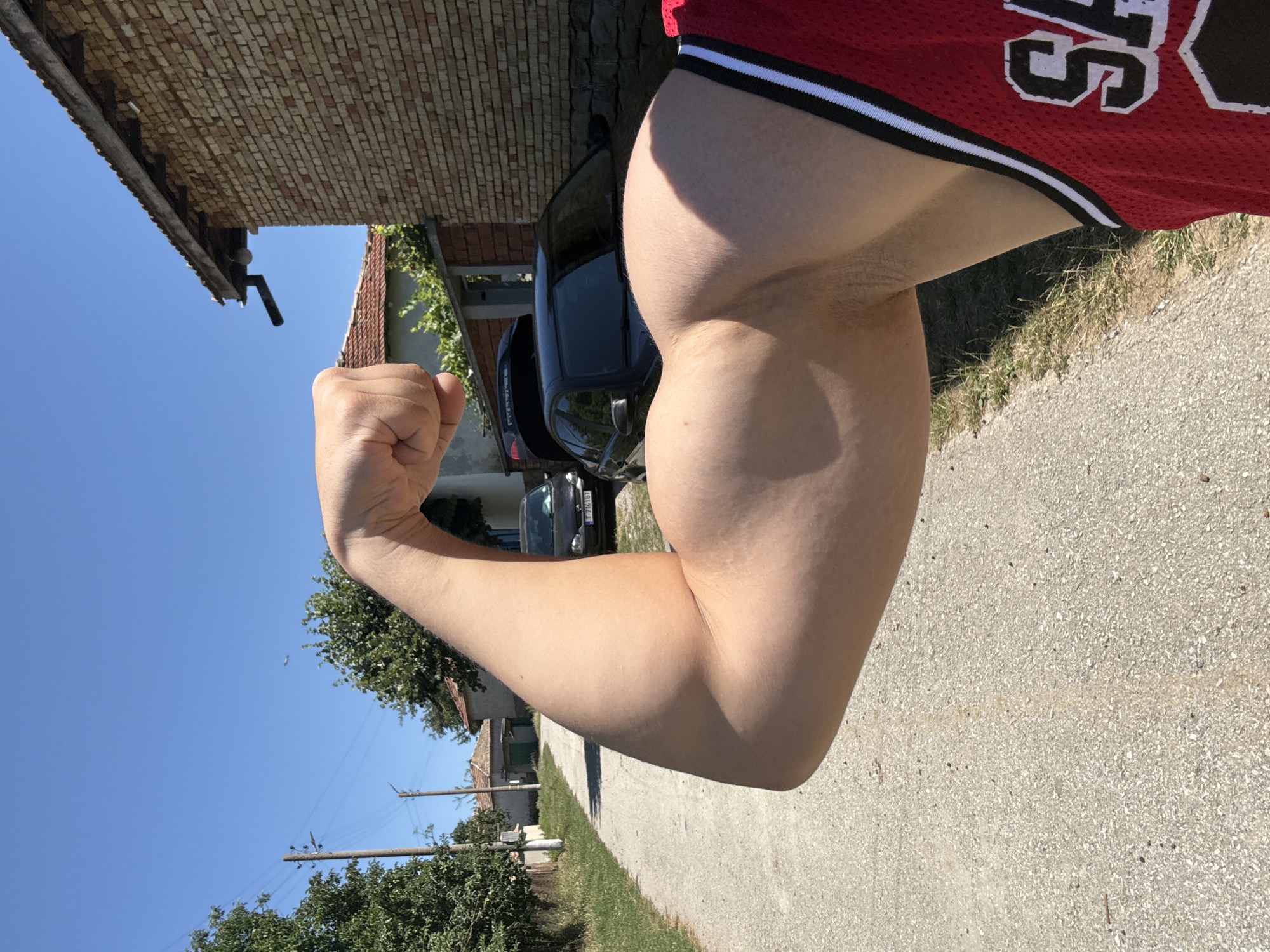 A person's flexed arm and bicep are prominently featured in the foreground. The individual is wearing a red basketball jersey with white and black trim, and the letters "SA" are visible on the jersey. The background depicts a sun-drenched rural street in the village of Konop, Bulgaria. A dirt road curves to the left, bordered by green grass and trees. Several houses with red-tiled roofs and brick facades are visible, as are two black cars parked on the right side of the road. A power line with poles stretches across the blue sky. The overall scene suggests a warm, clear day in a village setting.