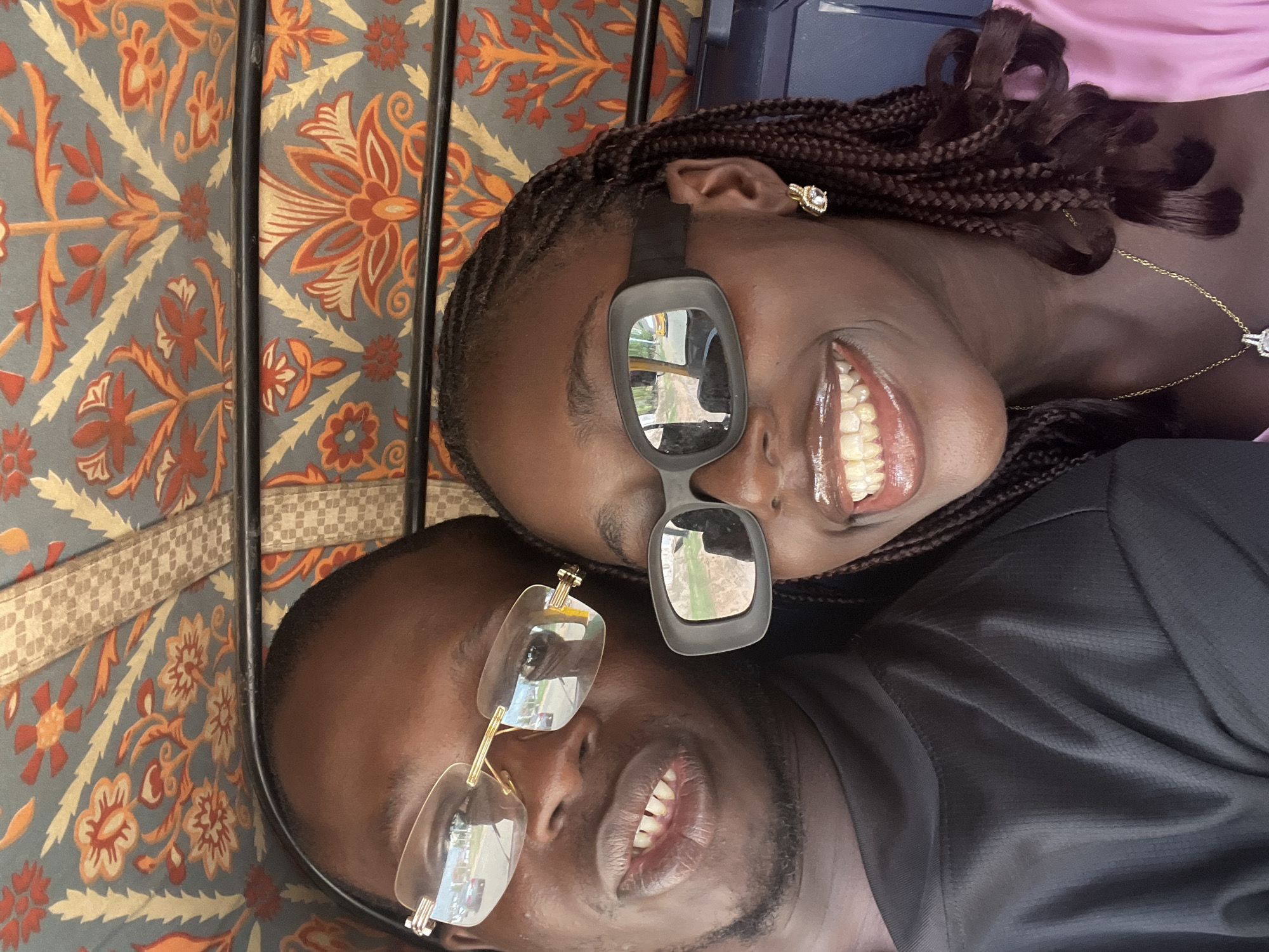 A close-up photo shows two individuals indoors. The individual on top is a woman with dark braided hair. She is wearing black sunglasses with rectangular lenses, showing reflections, and a pink top. She has an earring in her left ear and a gold necklace. The person below her is a man with short dark hair. He is wearing transparent glasses with gold frames and a black shirt. They both are smiling. The background has an intricate patterned fabric, with hues of grey, orange, and gold. There is a black metal frame behind the fabric. The scene appears to be inside a vehicle, possibly a keke (tricycle taxi), based on the background fabric and the angle. The location could be in Abuja, Nigeria, if that aligns with context and knowledge.