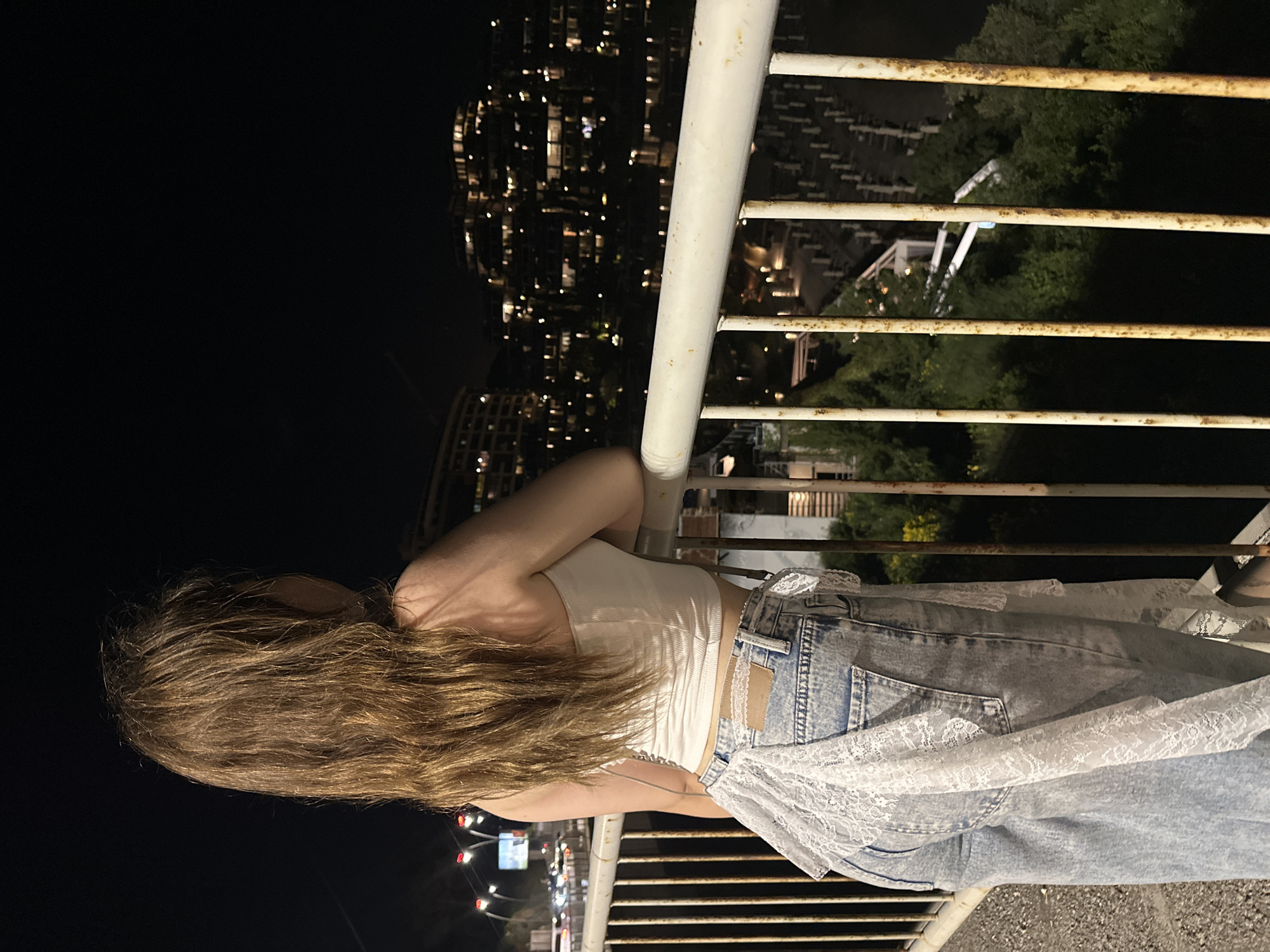 A woman with long, light brown hair is leaning on a white metal railing. She wears a white tank top and light wash jeans, with a white lace garment tied around her waist. The scene is set at night, with a cityscape visible in the background. The city is Budva, Montenegro, indicated by the distinctive cluster of tall, lit buildings in the background. Trees are present behind a secondary railing further away from the woman. The area beneath the railing shows a paved surface.