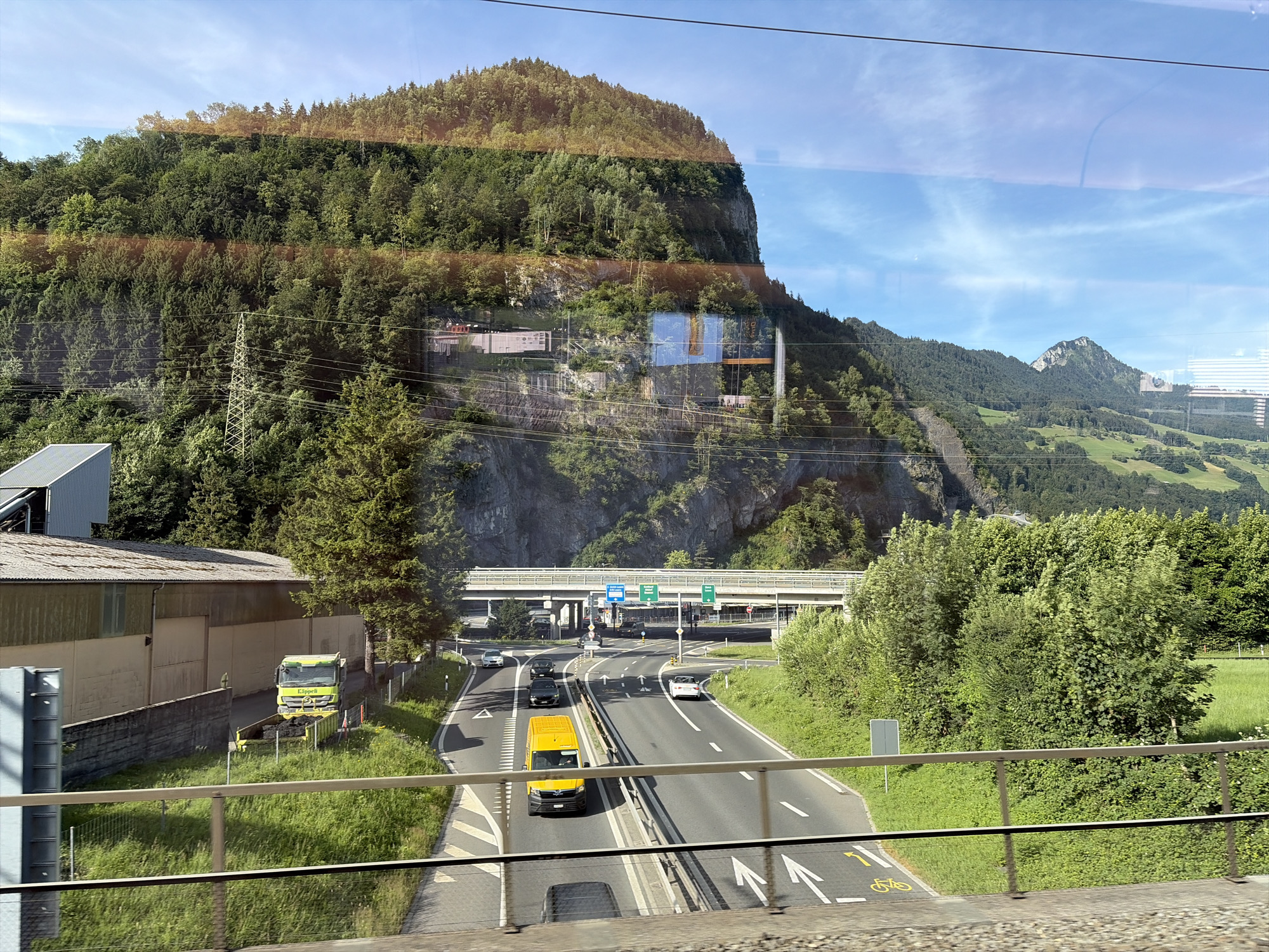 This image captures a view from a moving vehicle, likely a train, traveling along a highway in a mountainous region. The highway, a multi-lane asphalt road, is populated by several vehicles including cars, a yellow van, and a yellow truck. The truck appears to be unloading a dark material into a yellow bin. The road is bordered by grassy verges and lush green trees. A pedestrian bridge crosses over the highway in the middle distance. In the background, a steep, tree-covered mountain rises dramatically, with some rock faces visible. The sky above is blue with scattered white clouds. Reflections from the vehicle's windows are visible throughout the image, distorting the scene slightly.
While the image provides a scenic overview of a mountainous landscape and a highway, it does not contain specific visual cues that definitively place the location within the city of Schwyz, Switzerland. The general topography suggests a location in the Swiss Alps, which would be consistent with Schwyz, but further information would be needed for precise identification.