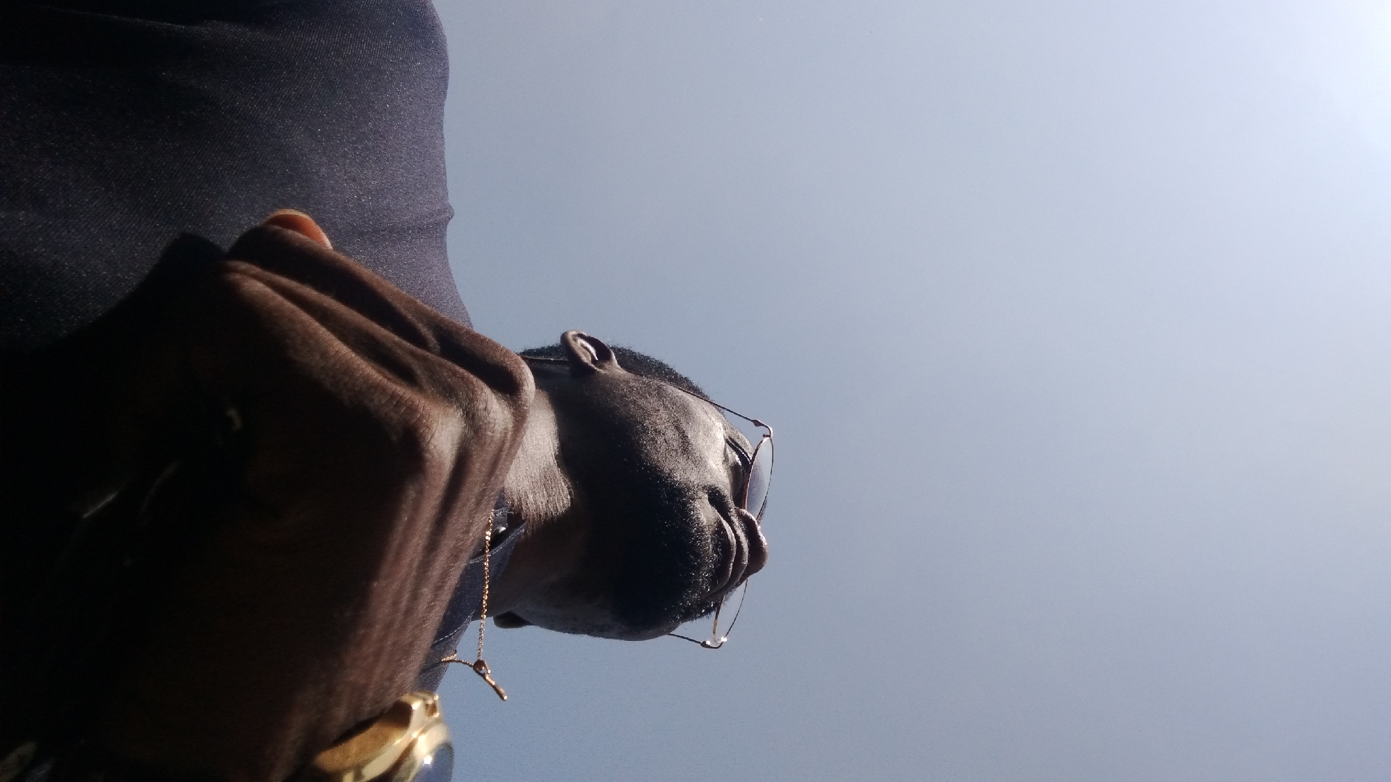 A low-angle, close-up, outdoor shot shows a Black man looking up into a clear, pale blue sky. He is wearing glasses with thin, metallic frames and a dark, textured shirt. A delicate gold chain is visible around his neck. His right hand is raised, partially obscuring his chest and shoulder, with his fingers loosely curled. He is wearing a gold watch on his left wrist. The lighting suggests it is daytime. The image does not contain any specific landmarks or clues that would allow for identification of a location within Jos, Nigeria.