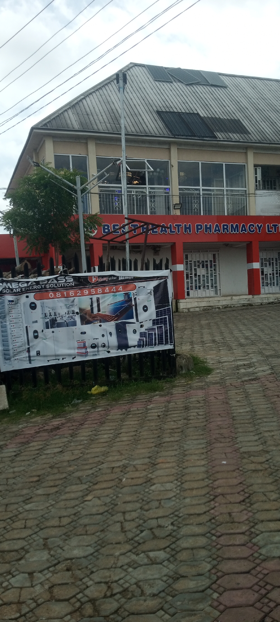 This media file shows the exterior of a two-story building with a red and white facade, identified by the text "BEST HEALTH PHARMACY LTD". The building has large windows on the upper floor, some of which are open, and solar panels are visible on the roof. A green tree is to the left of the entrance. In front of the building, a banner advertises "MEGA GLASS SOLAR ENERGY SOLUTION" and displays a phone number: "08162958444". The ground in front of the building and banner is paved with interlocking bricks. The sky is overcast, suggesting cloudy weather. The overall scene appears to be a street view in Jalingo, Nigeria, likely during daytime.