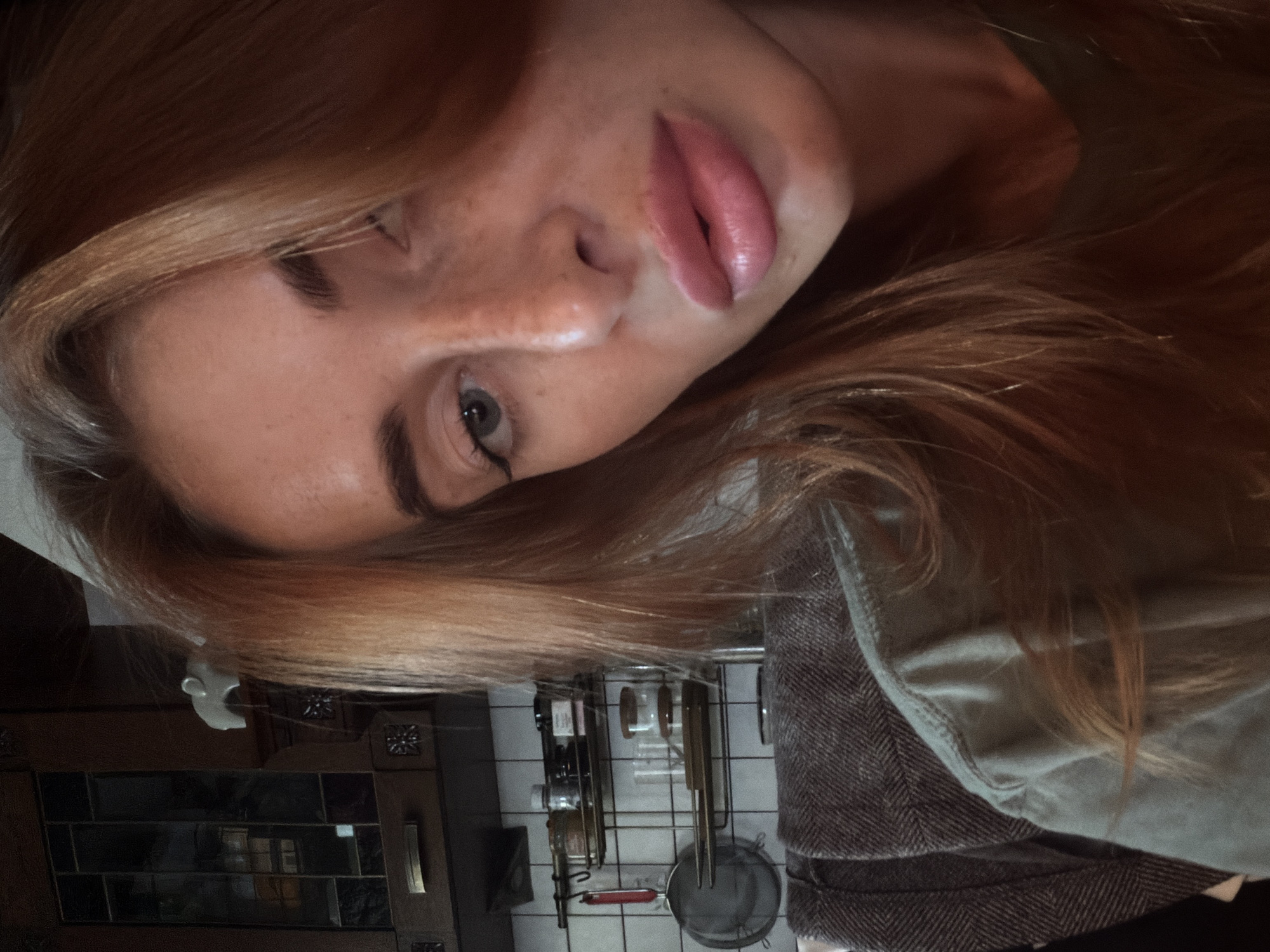 A selfie of a fair-skinned woman with red hair and freckles. She is wearing lipstick and gazing at the camera. Behind her is a kitchen scene with white tiled walls and various utensils hanging on a rack. There is also a dark wooden cabinet with glass panels and patterned borders. The location is identified as a kitchen, but specific details about its location in Krasnoyarsk, Russia are not definitively stated or visible in the image.