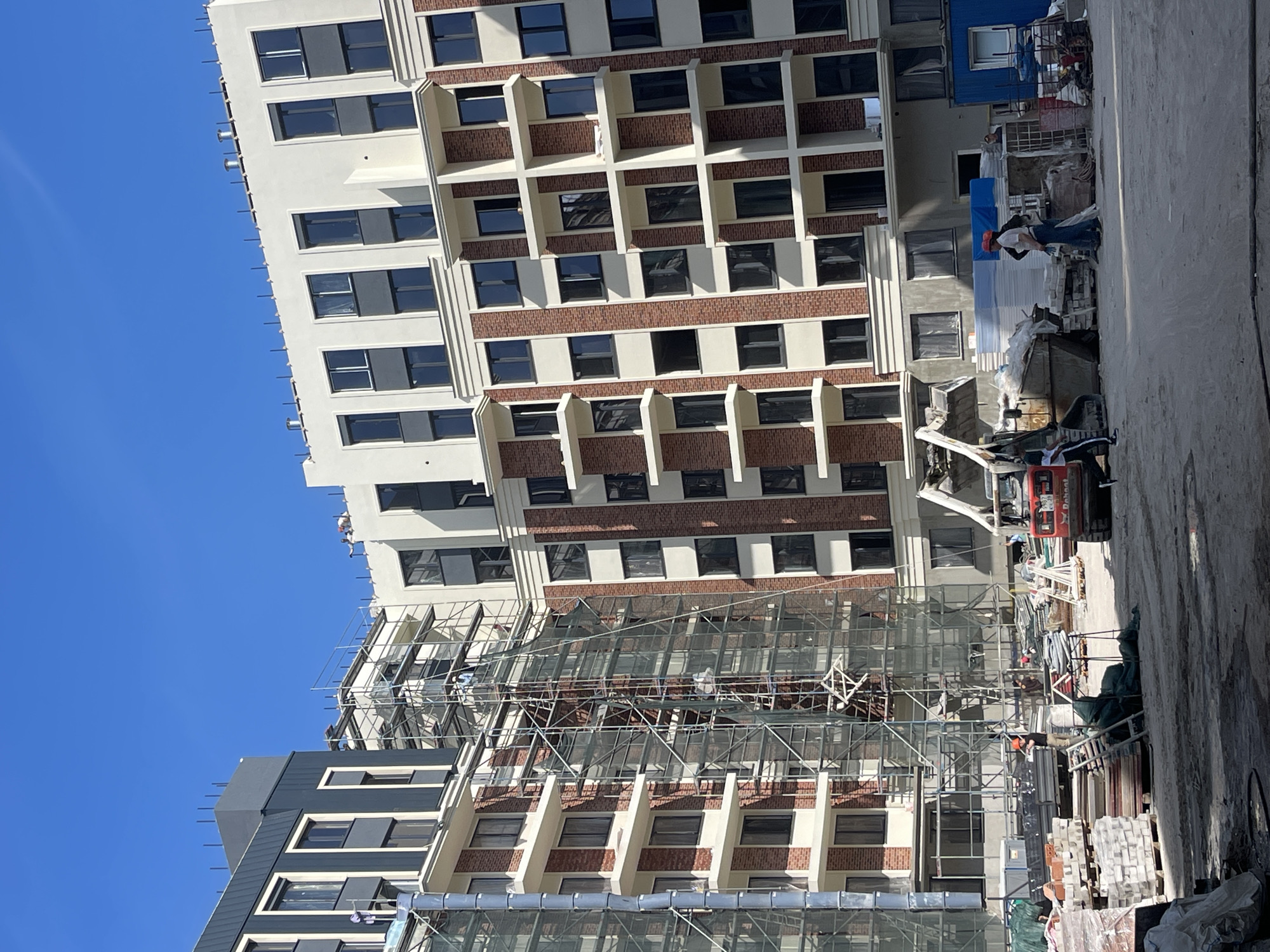 The image depicts a construction site in Zelenogradsk, Russia, focused on the exterior of a multi-story residential building under construction. The building features a modern design with a combination of white, grey, and red brick facades, accented by numerous dark-paned windows and white balconies. Extensive scaffolding covers a significant portion of the building's facade, indicating ongoing work.
In the foreground, the ground is a mix of dirt and sand, typical of a construction environment. Several stacks of building materials, including what appear to be bricks, plasterboard, and wooden planks, are visible. A red Bobcat mini-excavator is positioned on the right side of the frame, its bucket raised. Adjacent to it, two individuals, likely construction workers, are present. One is wearing a white shirt and blue jeans, and the other is wearing a dark shirt and blue jeans. They appear to be interacting or observing the ongoing work. Further to the left, another person in a dark shirt and blue jeans is standing near a pile of materials.
In the middle ground, several construction workers are visible on the scaffolding. One worker in a blue shirt is positioned higher up on the scaffolding, seemingly engaged in work. Another worker in a white shirt and orange helmet is near the Bobcat, facing away from the camera and holding what might be a tool. In the background, the clear blue sky serves as the backdrop to the partially completed building. The overall scene is one of active construction with workers and machinery present on site.