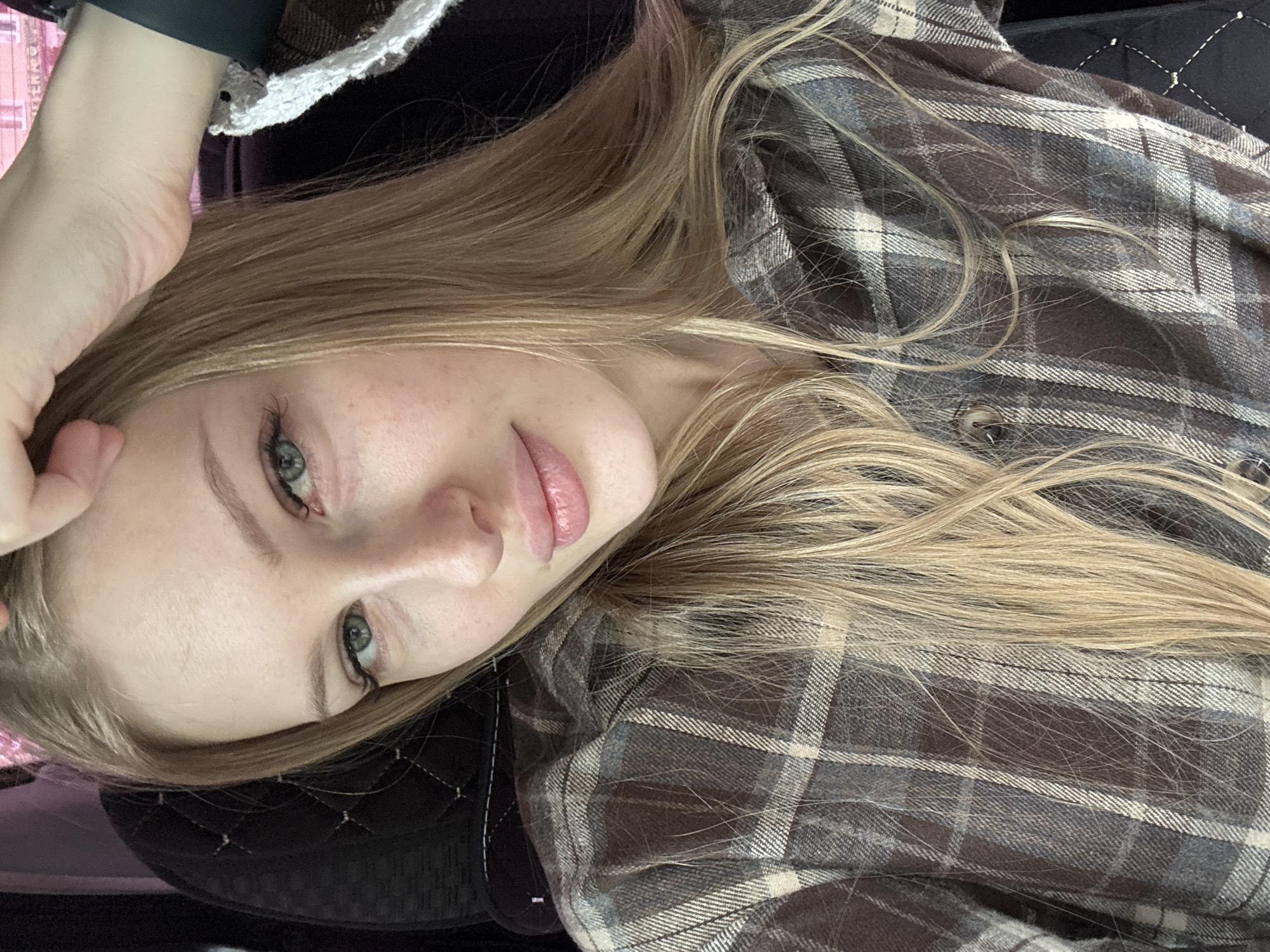 A medium close-up, canted view shows a woman lying down, her head resting on her left hand. She has fair skin, light blonde hair, and light-colored eyes. She is wearing a brown and grey plaid shirt. The background includes a quilted black material and some ambiguous colors, but no specific location details are discernible in the image. The location in Perm, Russia is not discernible from the image.