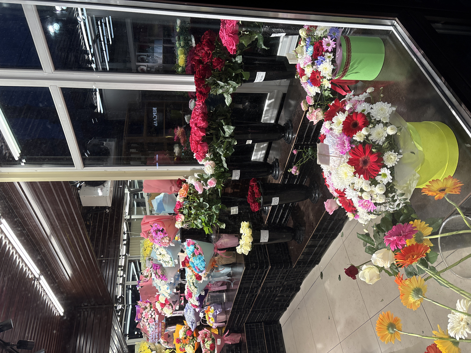 This image captures a vibrant and extensive display of flowers within what appears to be a flower shop, viewed through a large glass window. The scene is brightly lit by artificial lights, casting reflections on the glass that suggest an evening or nighttime setting outside.
Numerous flower arrangements are visible, showcasing a wide variety of blooms and colors. On the left side of the display, an array of bouquets, wrapped in colorful paper and cellophane, are arranged on tiered shelving units. These include various types of flowers such as chrysanthemums, roses, and mixed assortments in shades of pink, purple, white, yellow, and blue.
Towards the center and right, large bundles of cut roses, predominantly red and pink, are presented in tall black vases. Further right, more elaborate mixed bouquets featuring gerberas, chrysanthemums, and roses are stylishly arranged in round green hat-boxes. On the floor in the foreground, several loose single-stem gerberas in vivid orange, yellow, pink, and white, along with a few white and red roses, are displayed.
The glass window reflects a dark exterior, along with the distinct shapes of fluorescent tube lights from the shop's interior ceiling. Part of a Cyrillic word, "БЕССКУР", is visible in reflection on the glass. Several price tags are also legible on the vases and bouquets, displaying prices such as "330.00", "300.00", "190", and "380", indicating that the flowers are for sale. The overall impression is one of a bustling and colorful floral retail environment.