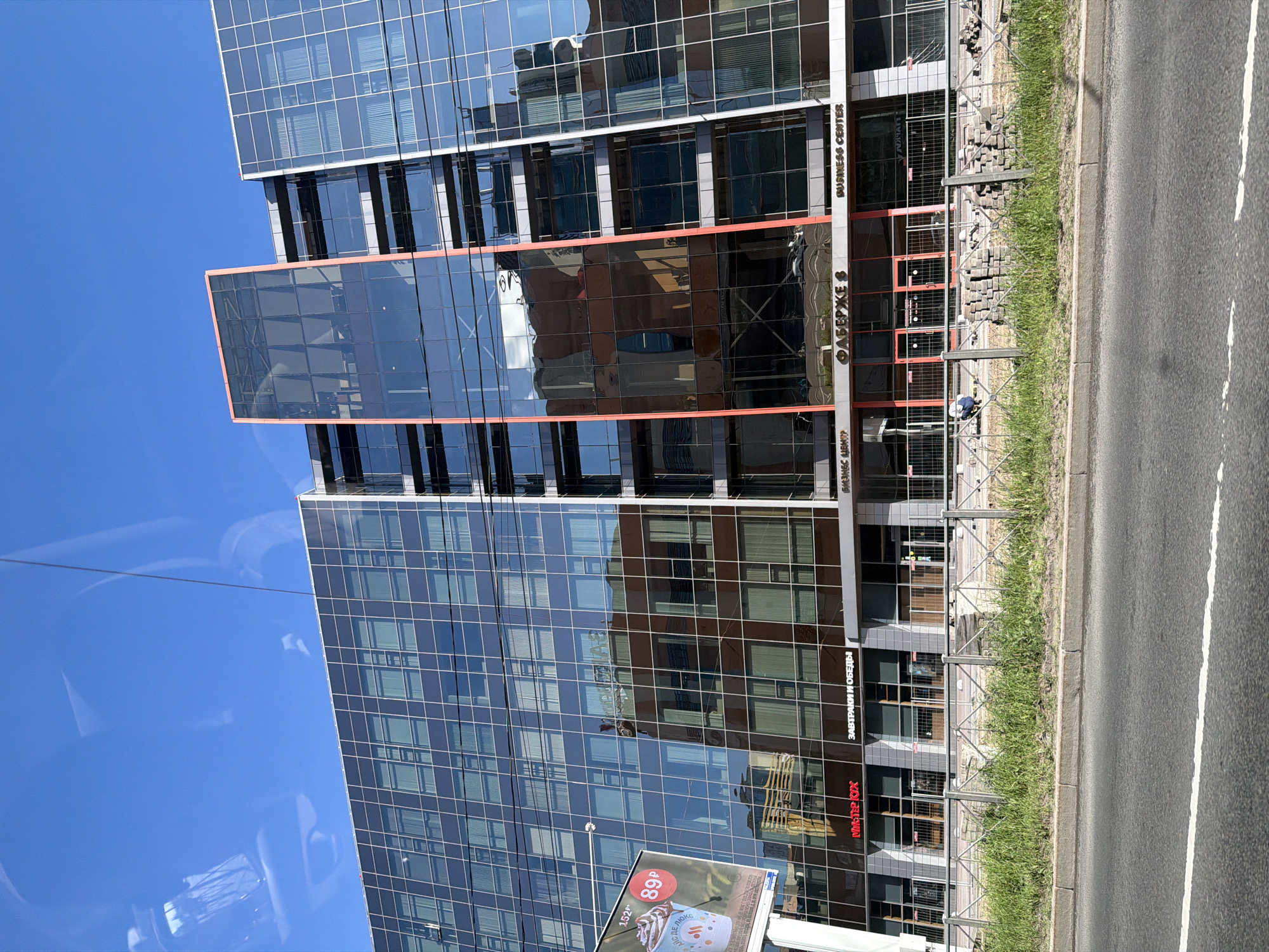 The image captures a modern, multi-story glass-facade business center, identified as "БИЗНЕС ЦЕНТР ФАБЕРЖЕ 8" (BUSINESS CENTER FABERGE 8), located at Koltushskoye Shosse, 44к1, Vsevolozhsk, Leningrad Oblast, Russia. The building features a prominent vertical section outlined in orange-red near its center, with the rest of the facade comprising dark-framed glass panels reflecting the clear blue sky and ambient surroundings.
The ground level of the building includes commercial spaces with signage: "Мистер Кух" (Mister Kukh) and "ЗАВТРАКИ И ОБЕДЫ" (Breakfasts and Lunches) on the left, and "ФАБЕРЖЕ 8" (FABERGE 8) and "BUSINESS CENTER" (in English) on the right. In front of these ground-level entrances, a metal fence separates the building from a strip of grass. Behind this fence, stacks of light-colored paving stones are neatly arranged, and a person in a blue shirt is visible crouching near these materials, suggesting ongoing construction or landscaping work.
To the far left of the building, a billboard displays an advertisement for a beverage, showing a crossed-out price of "152P" and a current price of "89P" in a red circle, with the text "ГДЕ ДЕ ЛЮКС" on the cup. In the immediate foreground, an asphalt road with a white dashed line is visible. The top portion of the image shows reflections consistent with being photographed from inside a vehicle, including faint outlines of a car's interior.