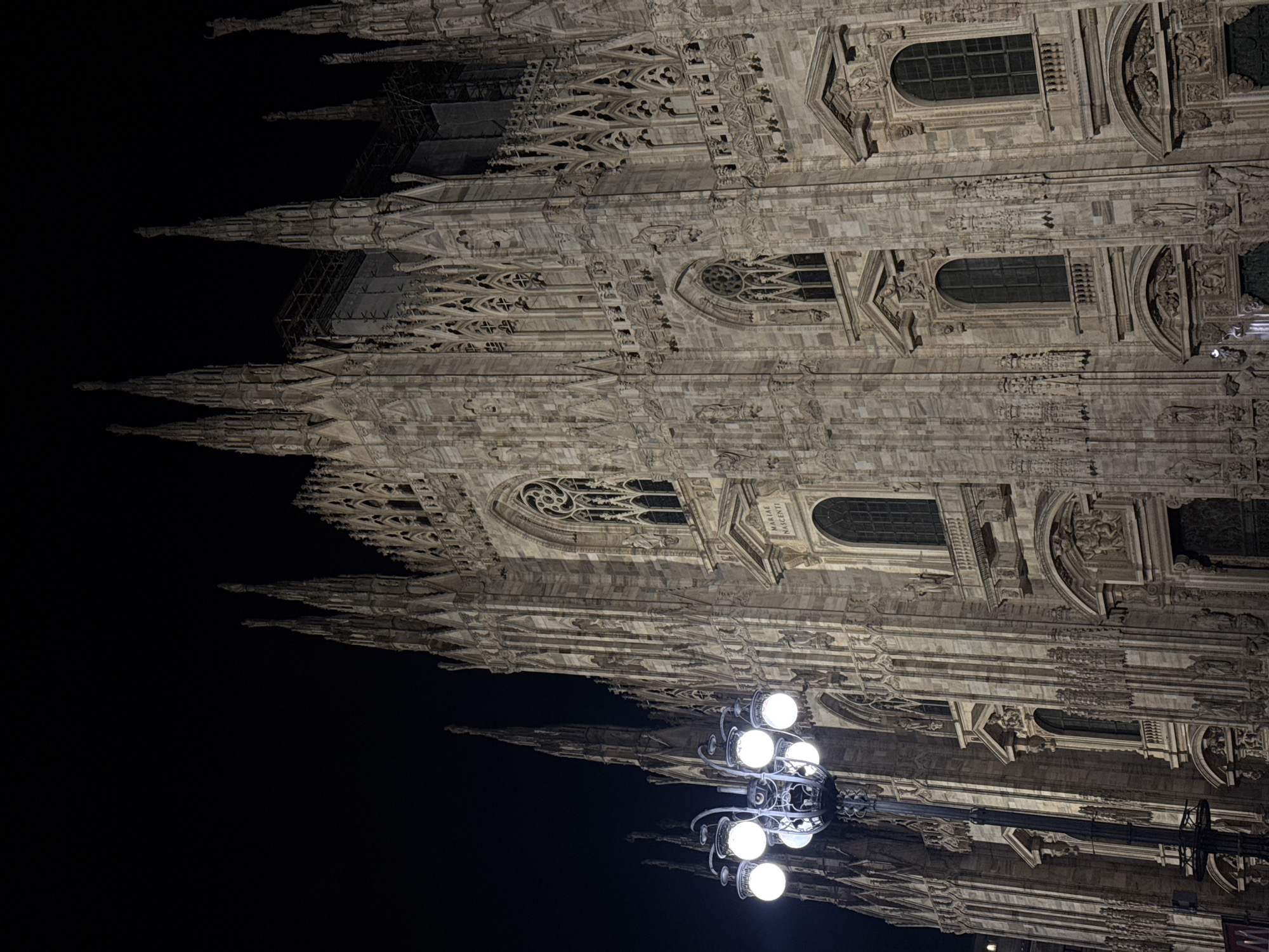 The image depicts an upward-facing, low-angle view of the Milan Cathedral (Duomo di Milano) at night. The illuminated Gothic architecture of the cathedral is contrasted against the dark night sky. A street lamp with multiple spherical bulbs is visible in the lower part of the frame.